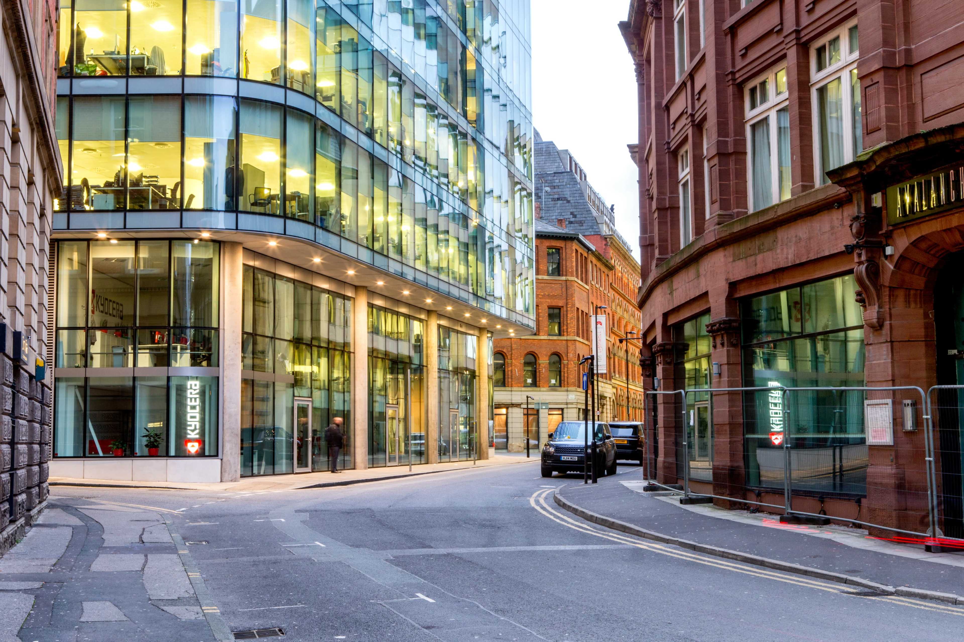 A modern glass building intersects with an older brick structure on a city street, where a van is parked near construction barriers.