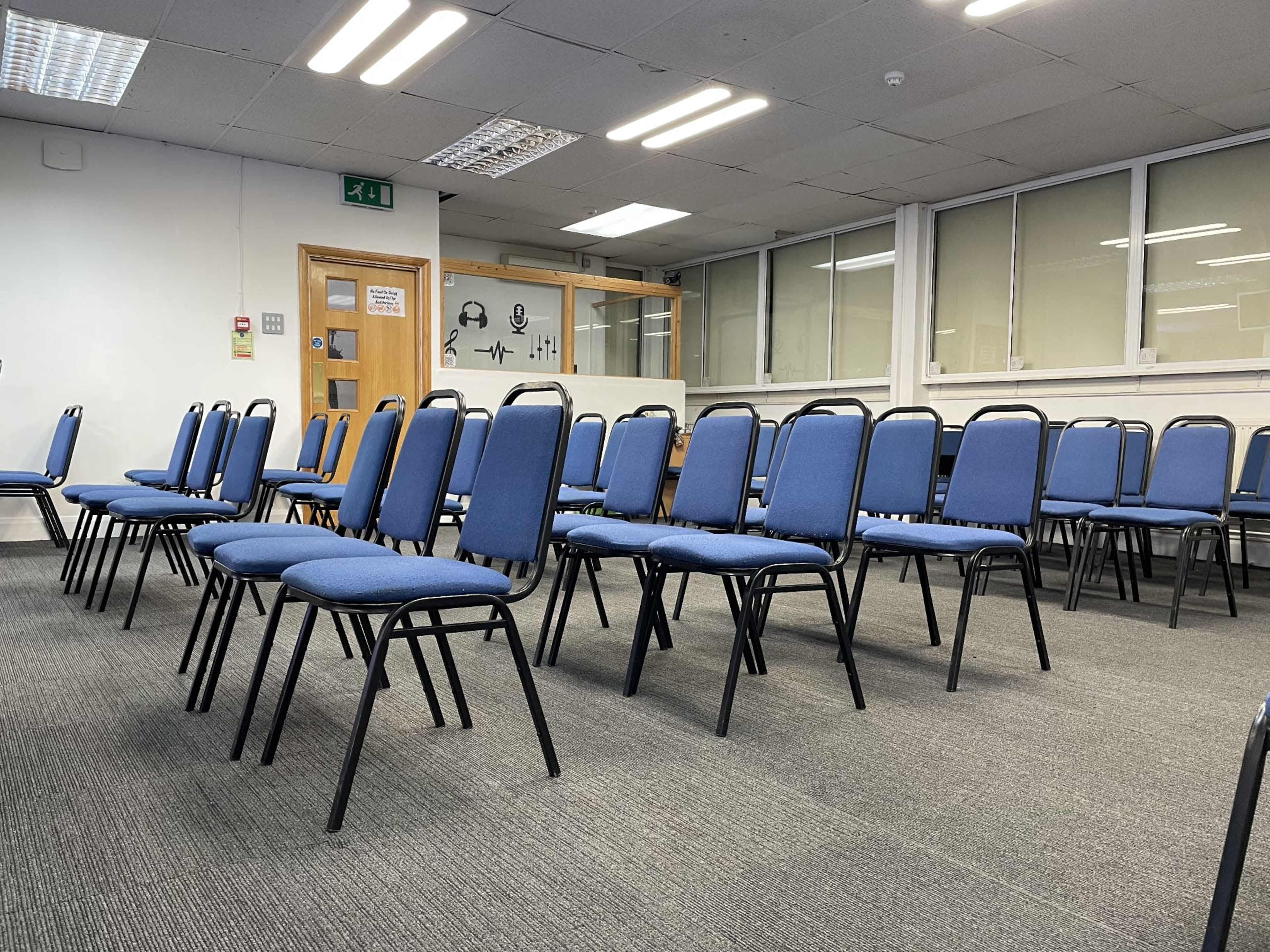 A row of blue chairs arranged in a room with carpeted flooring and large windows in the background.
