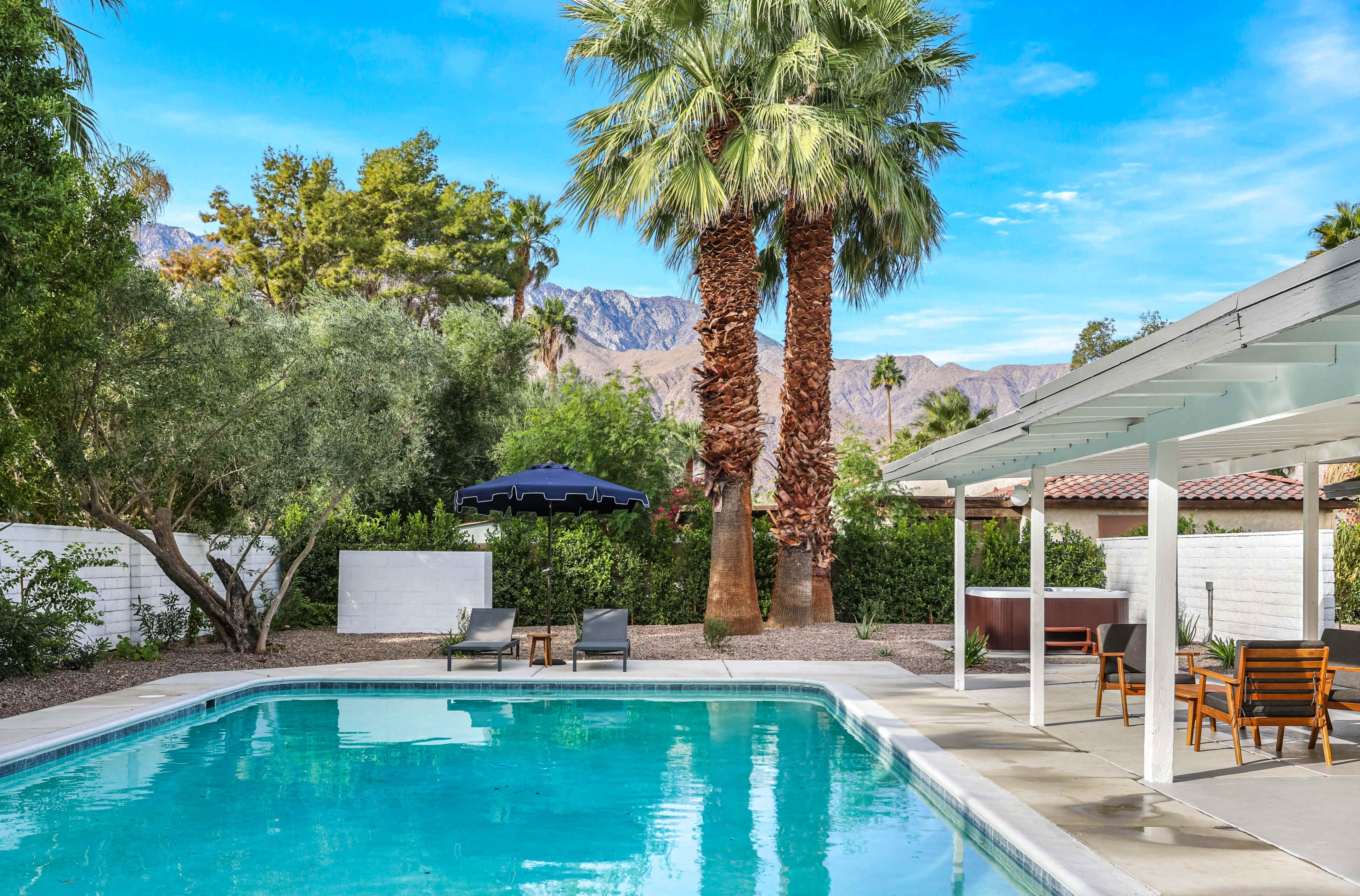 The image shows a clear swimming pool surrounded by palm trees and lounge chairs, with a backdrop of mountains under a blue sky.