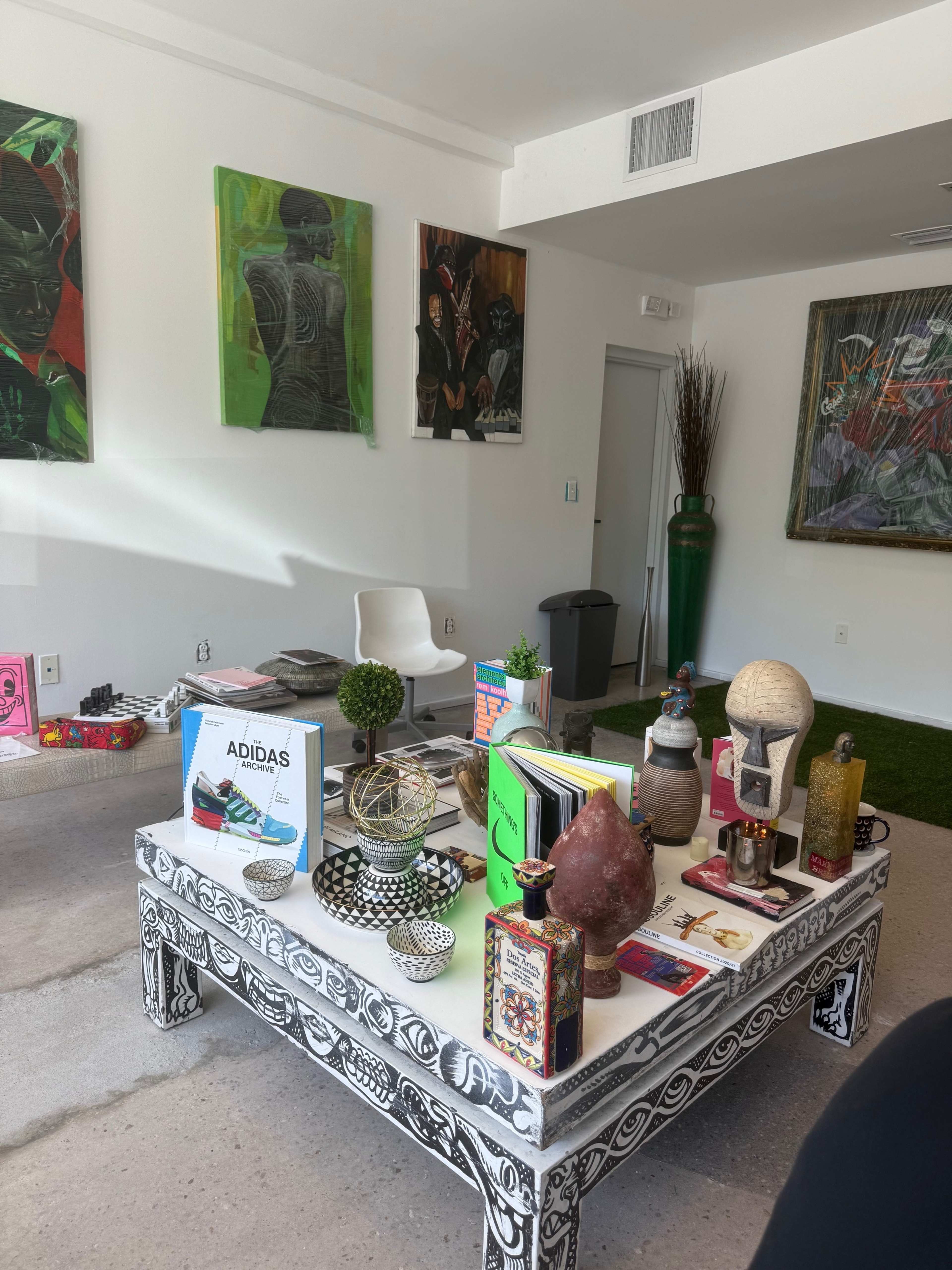 The image depicts a modern, well-lit room featuring a low coffee table adorned with various decorative items, books, and plants, surrounded by minimalist furniture and colorful artwork on the walls.