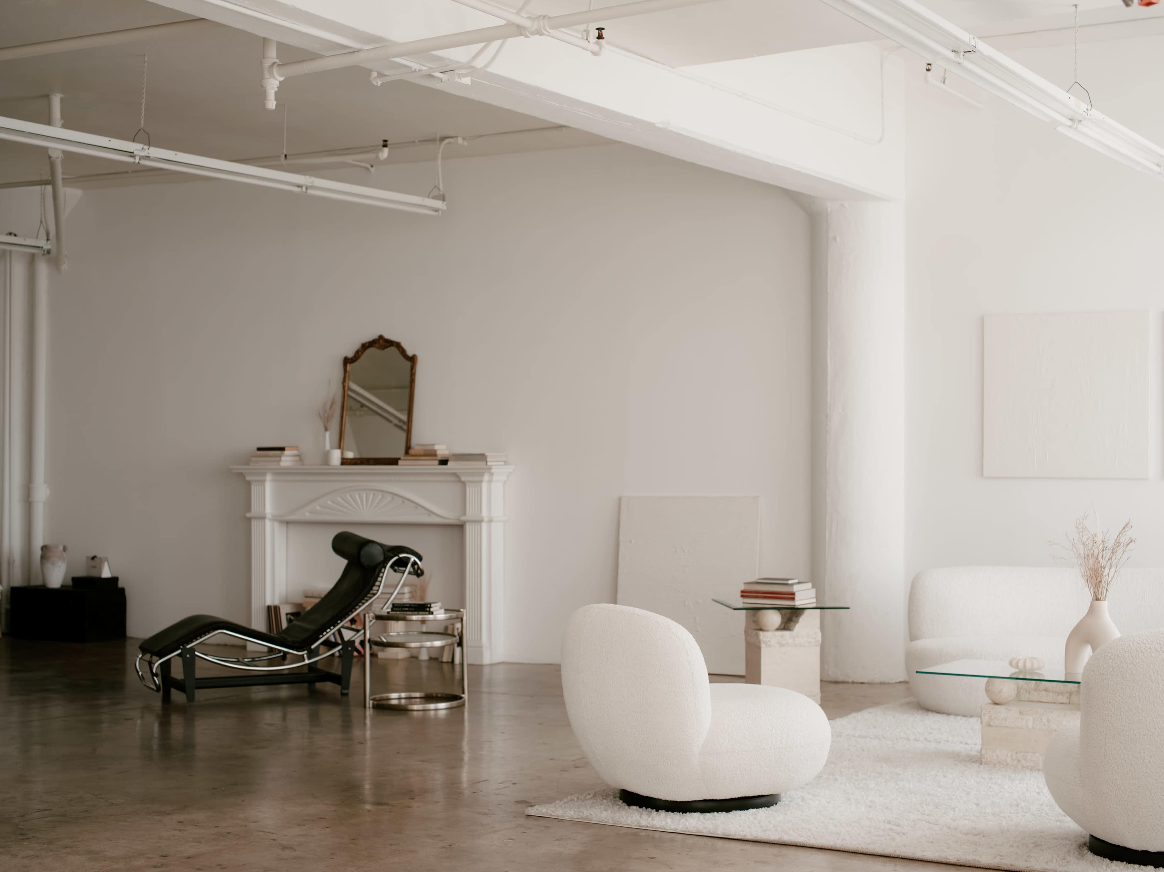 The image shows a minimalist interior space featuring a lounge area with white plush chairs, a glass coffee table, and a sleek chaise lounge, along with a decorative mirror above a mantel.