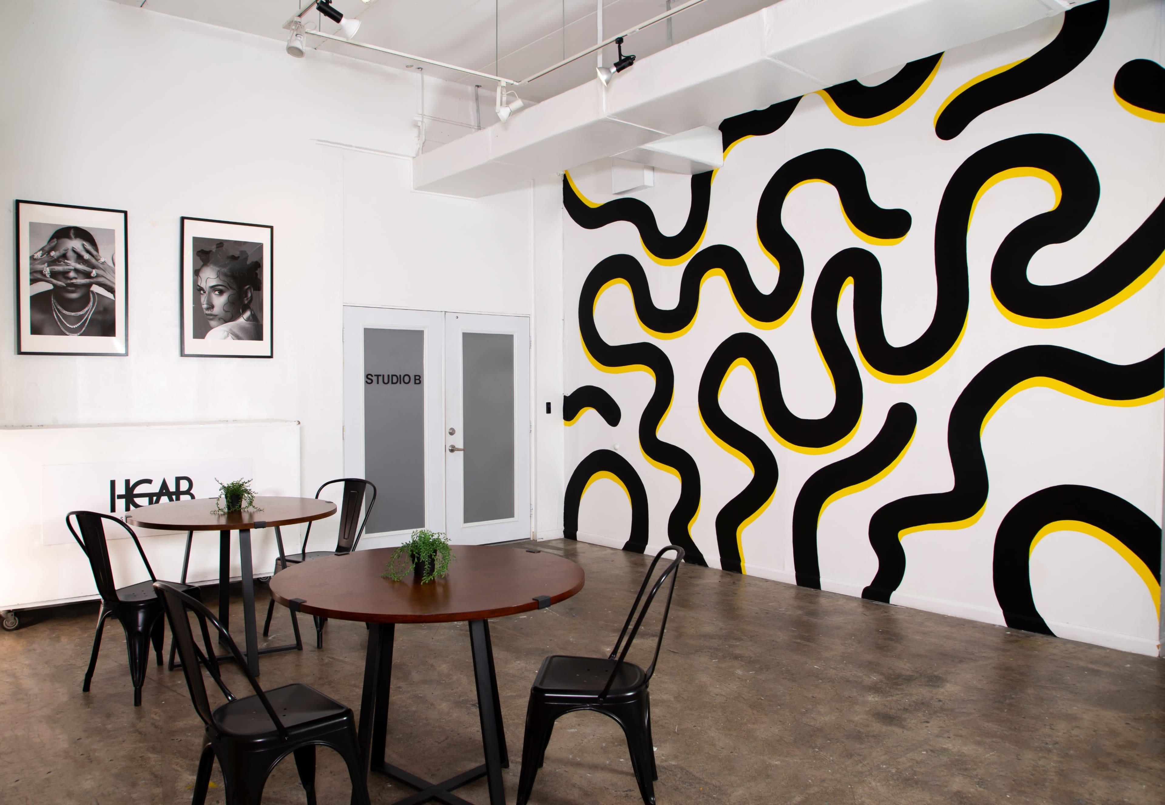 A modern studio space featuring a white wall with a black and yellow wave pattern, two framed photographs, and two round tables with black chairs.