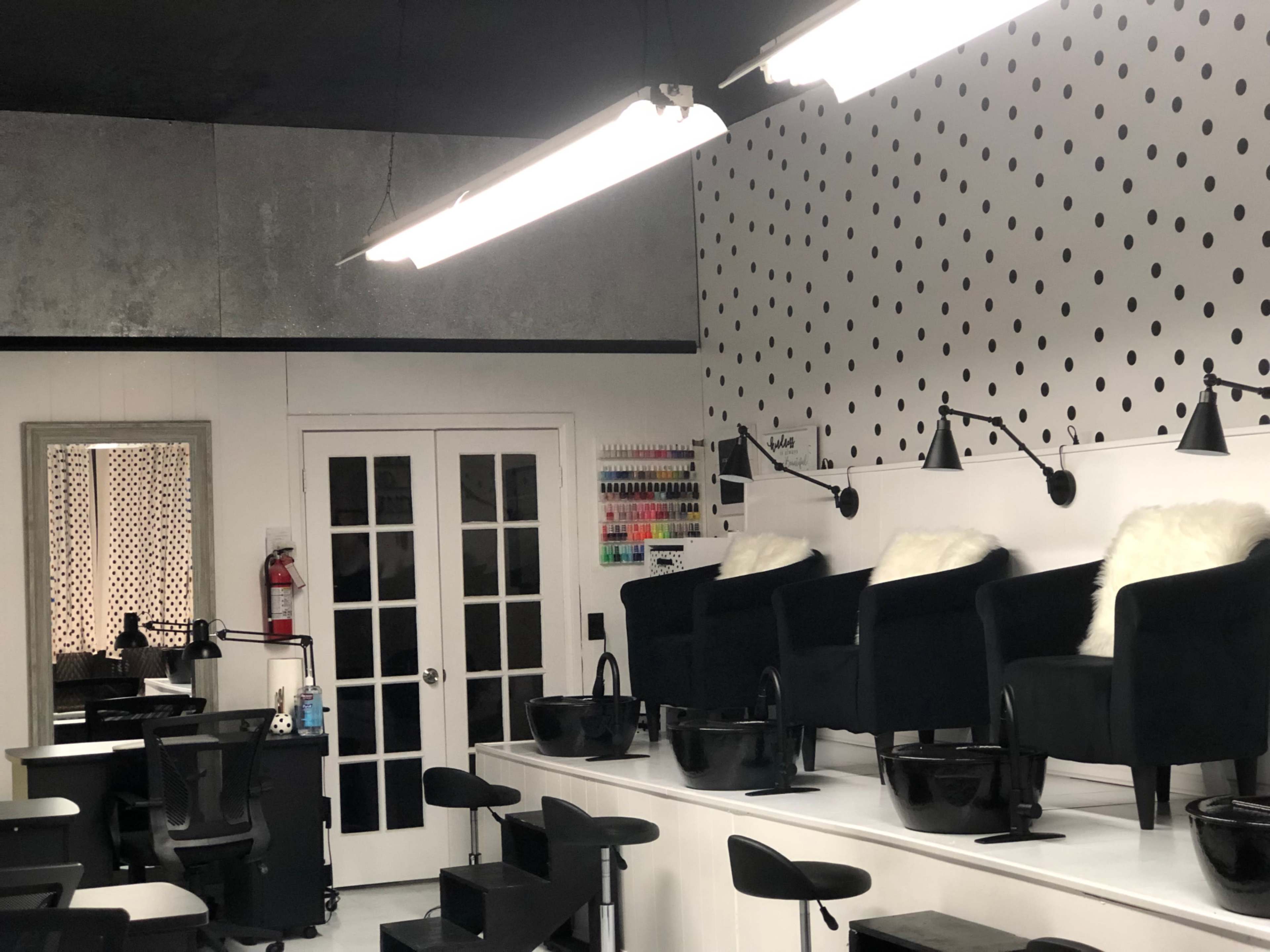 A nail salon interior featuring black chairs with white fur throws, nail polish displayed on a wall, and white walls adorned with black polka dots.