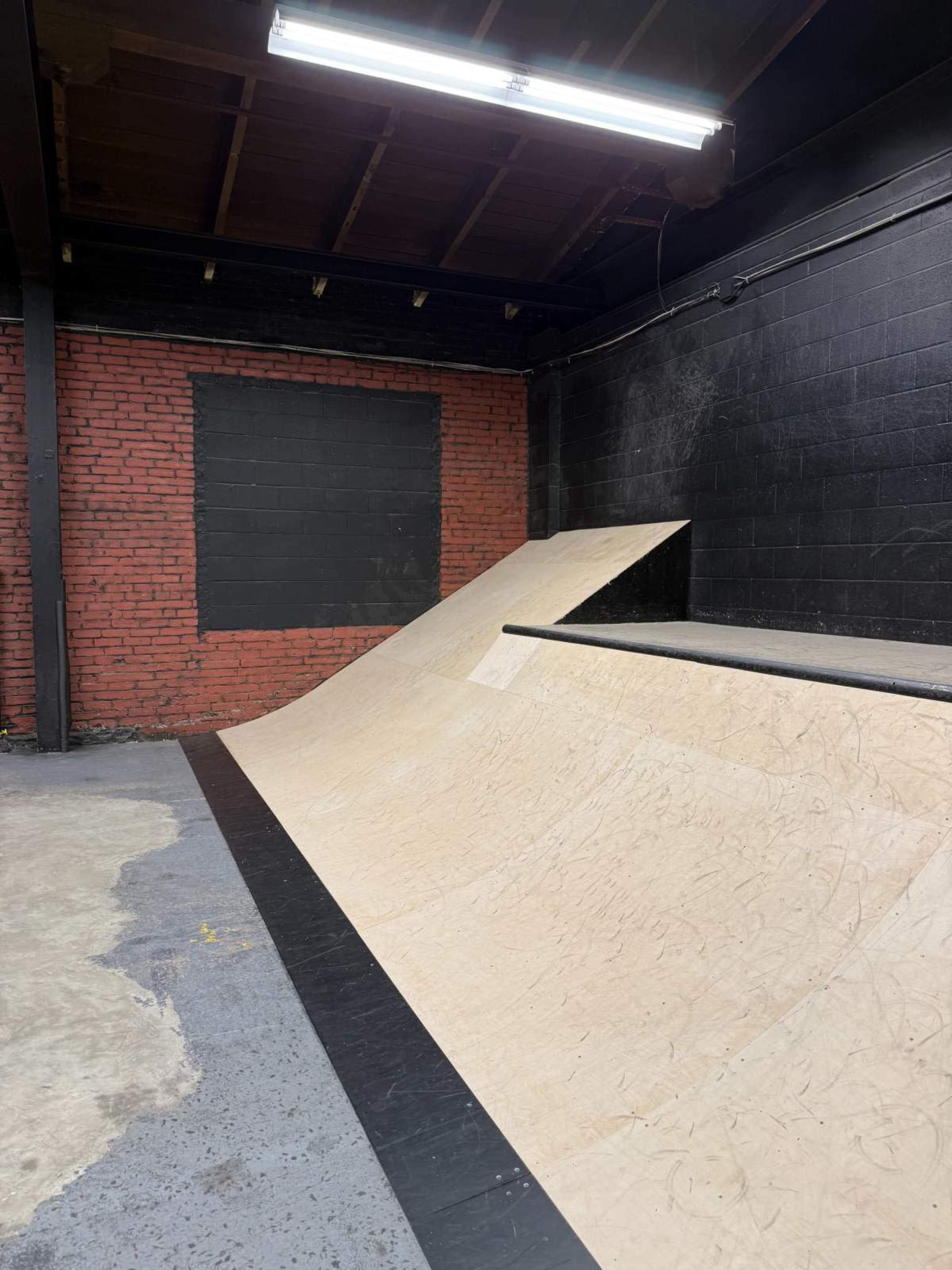 The image shows a wooden skate ramp inside a dimly lit indoor skate park with red brick and black walls.