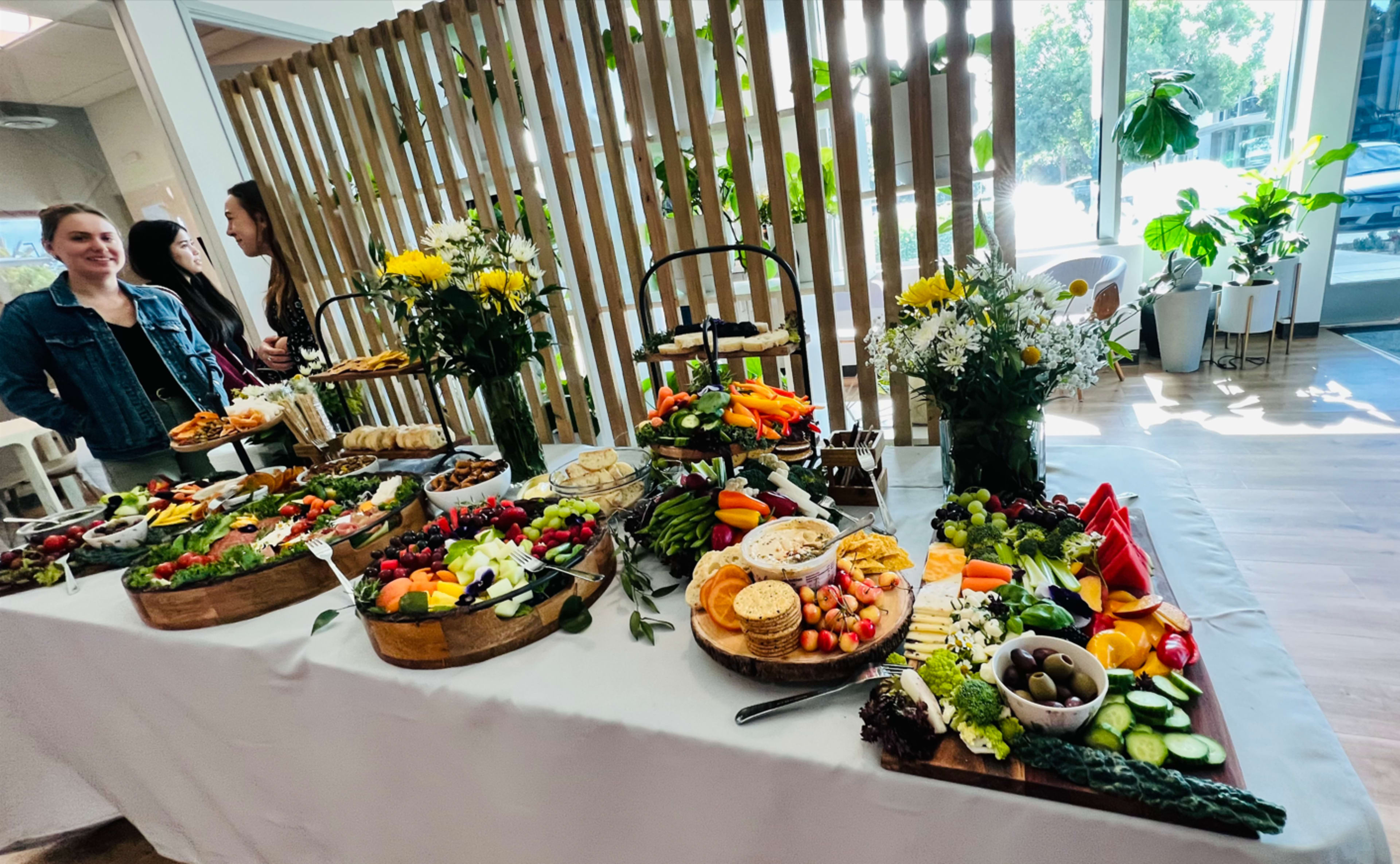 A rectangular table displays an array of colorful food items, including fruits, vegetables, cheeses, and crackers, with floral arrangements in the background.