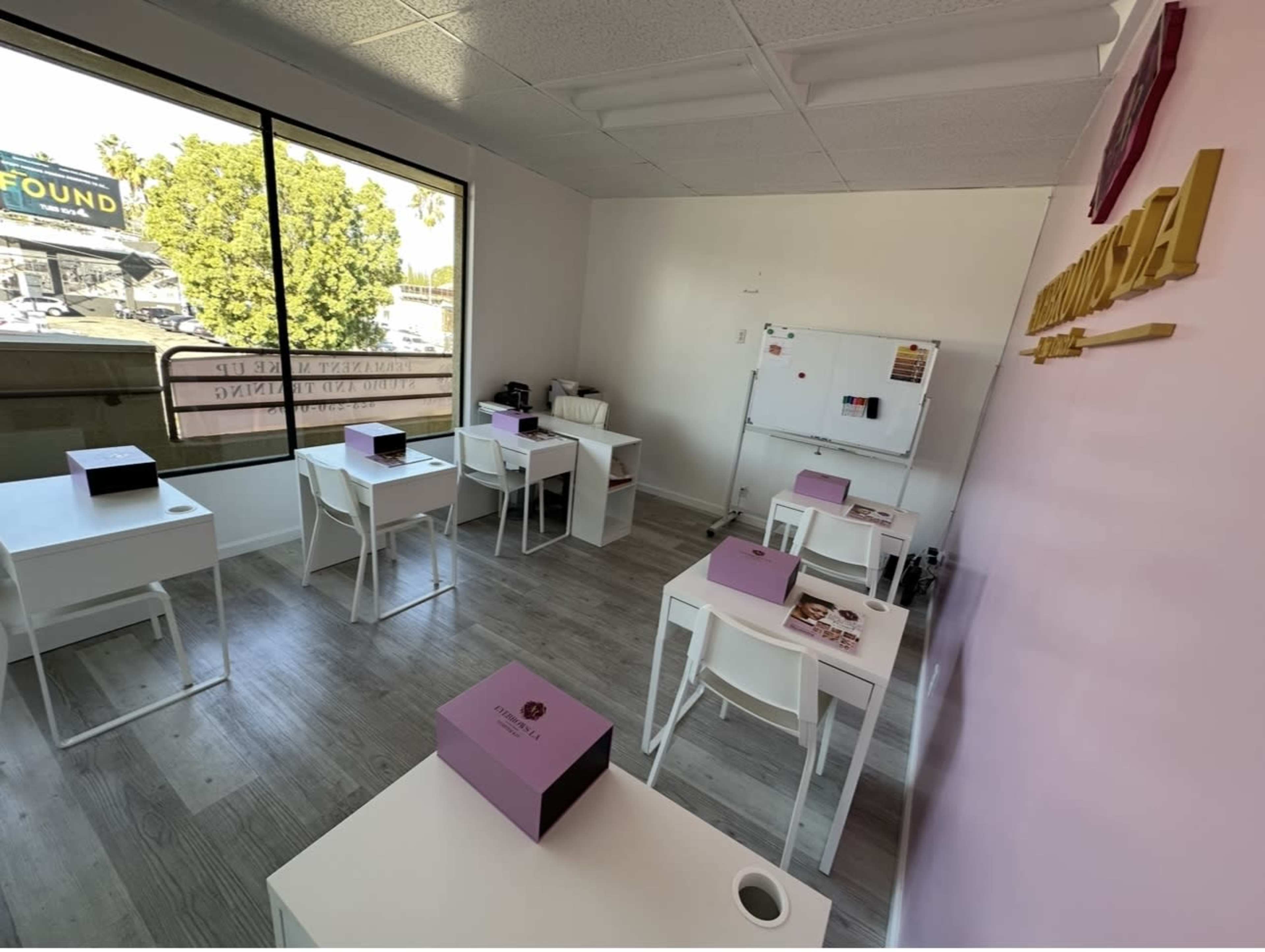 The image shows a light-colored classroom with multiple white desks, some with pink boxes on them, and a large window allowing natural light to enter.
