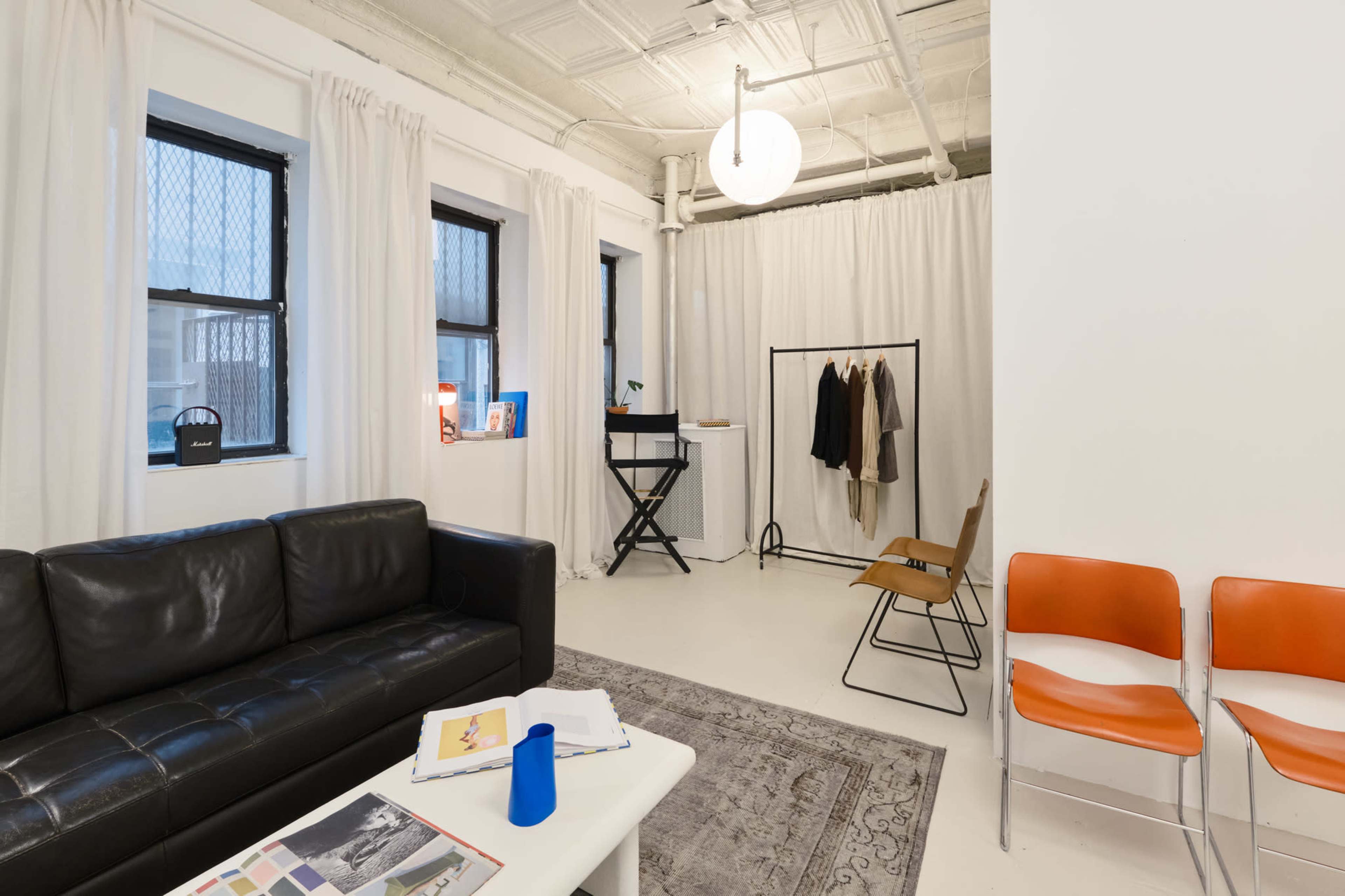 The image shows a modern, minimalist indoor space featuring a black leather sofa, a clothing rack with garments, a small desk, and orange chairs arranged around a light-colored rug.
