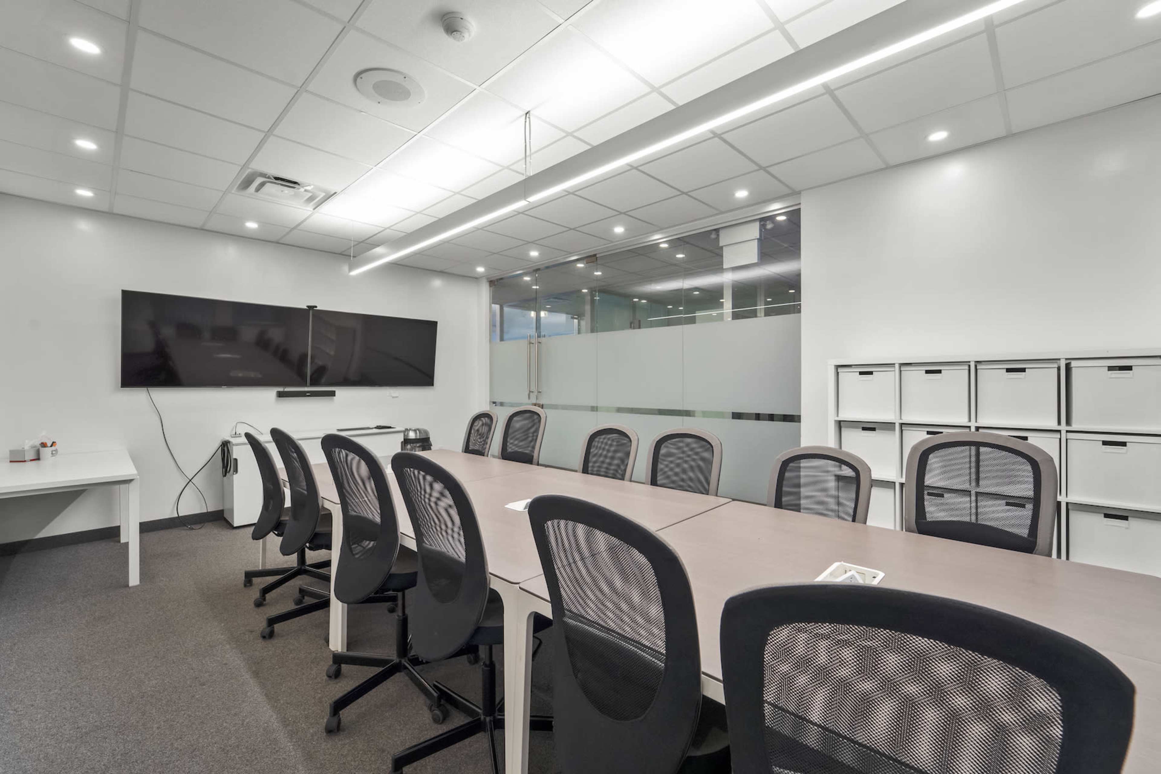 A modern conference room features a long table surrounded by multiple ergonomic chairs, a large screen mounted on the wall, and a glass partition providing a view of an adjacent space.