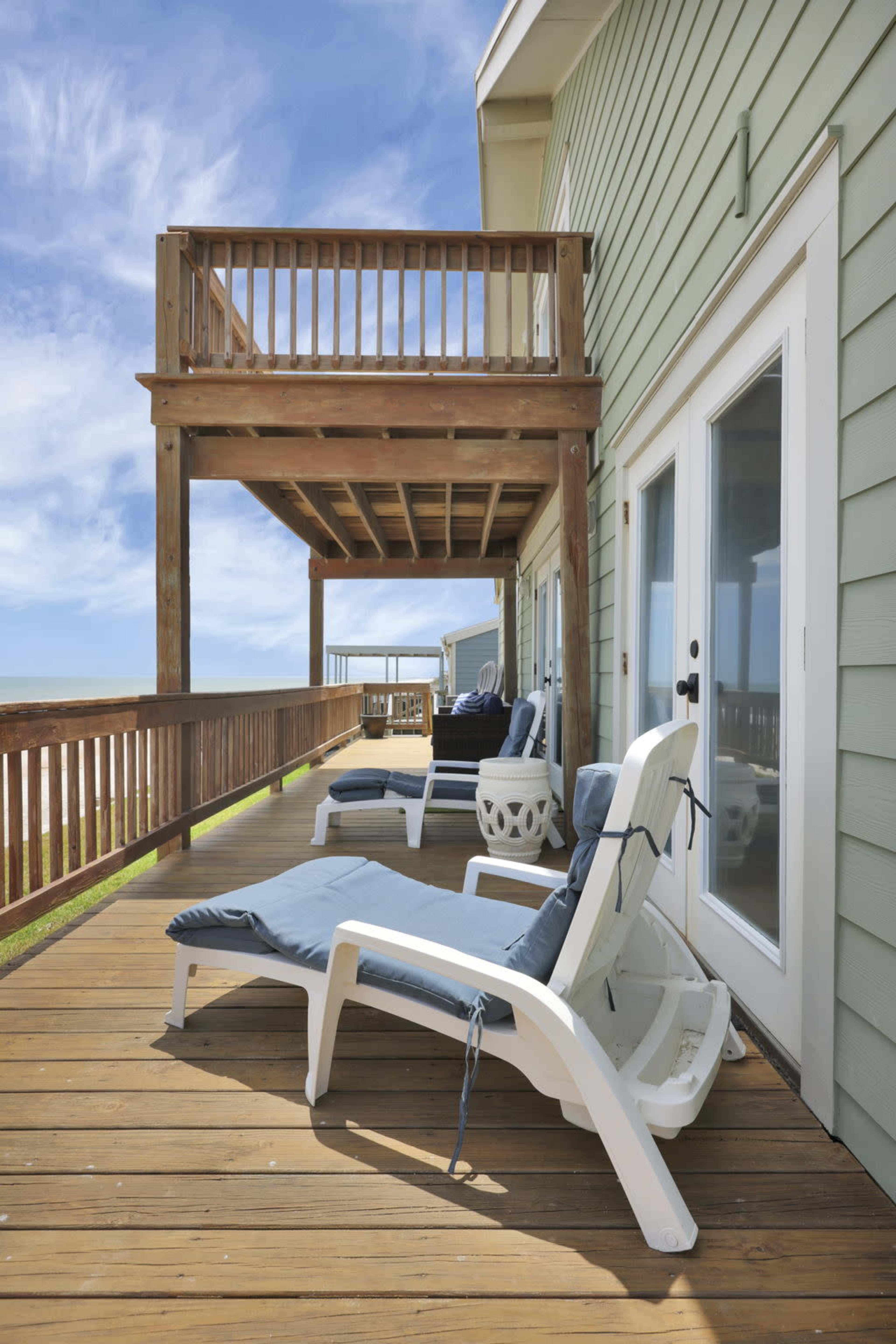 A wooden deck with lounge chairs and a second-story balcony attached to a light green house, overlooking a clear blue sky.