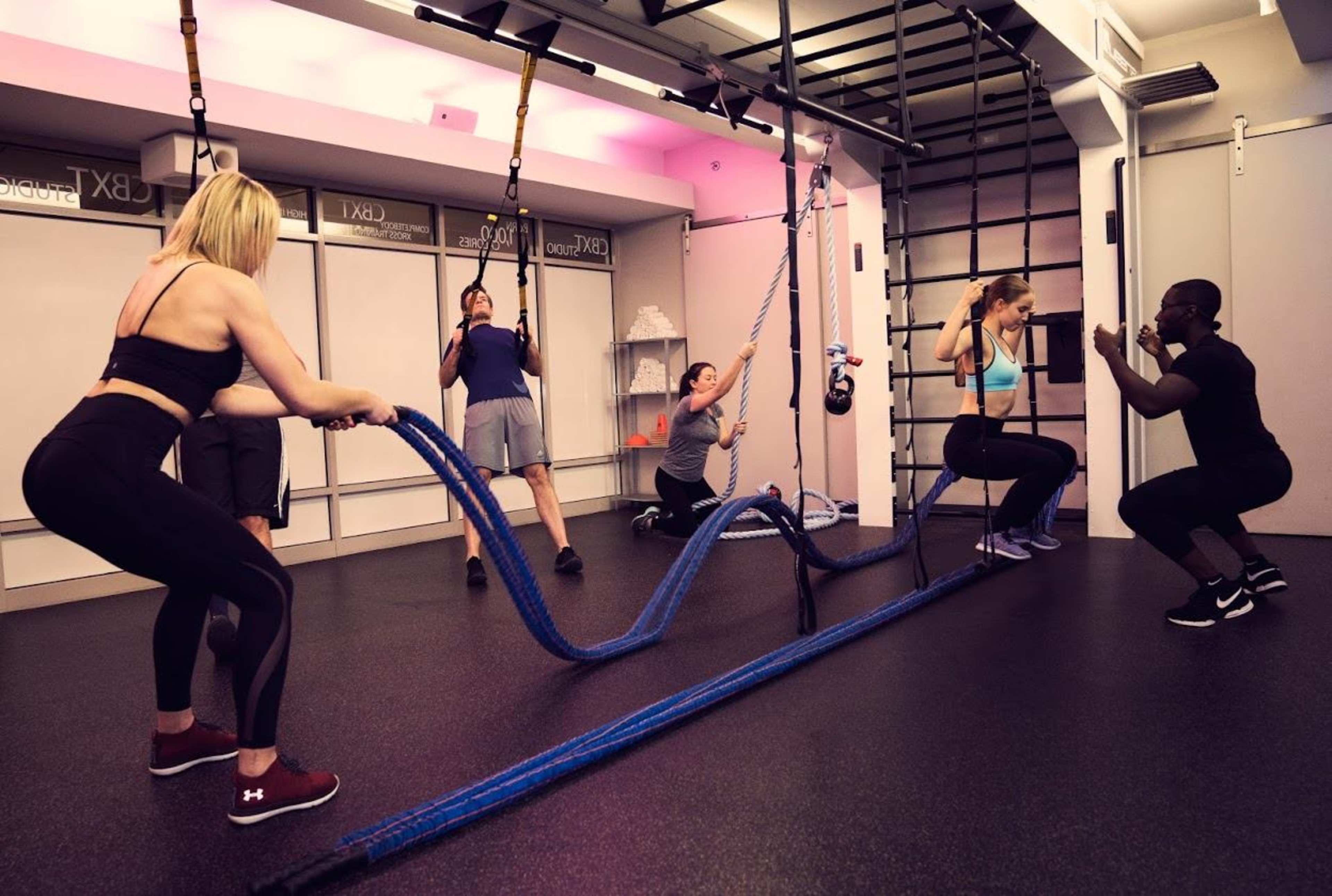 A group of people are exercising in a gym, using battle ropes, kettlebells, and performing various workouts on gym equipment.