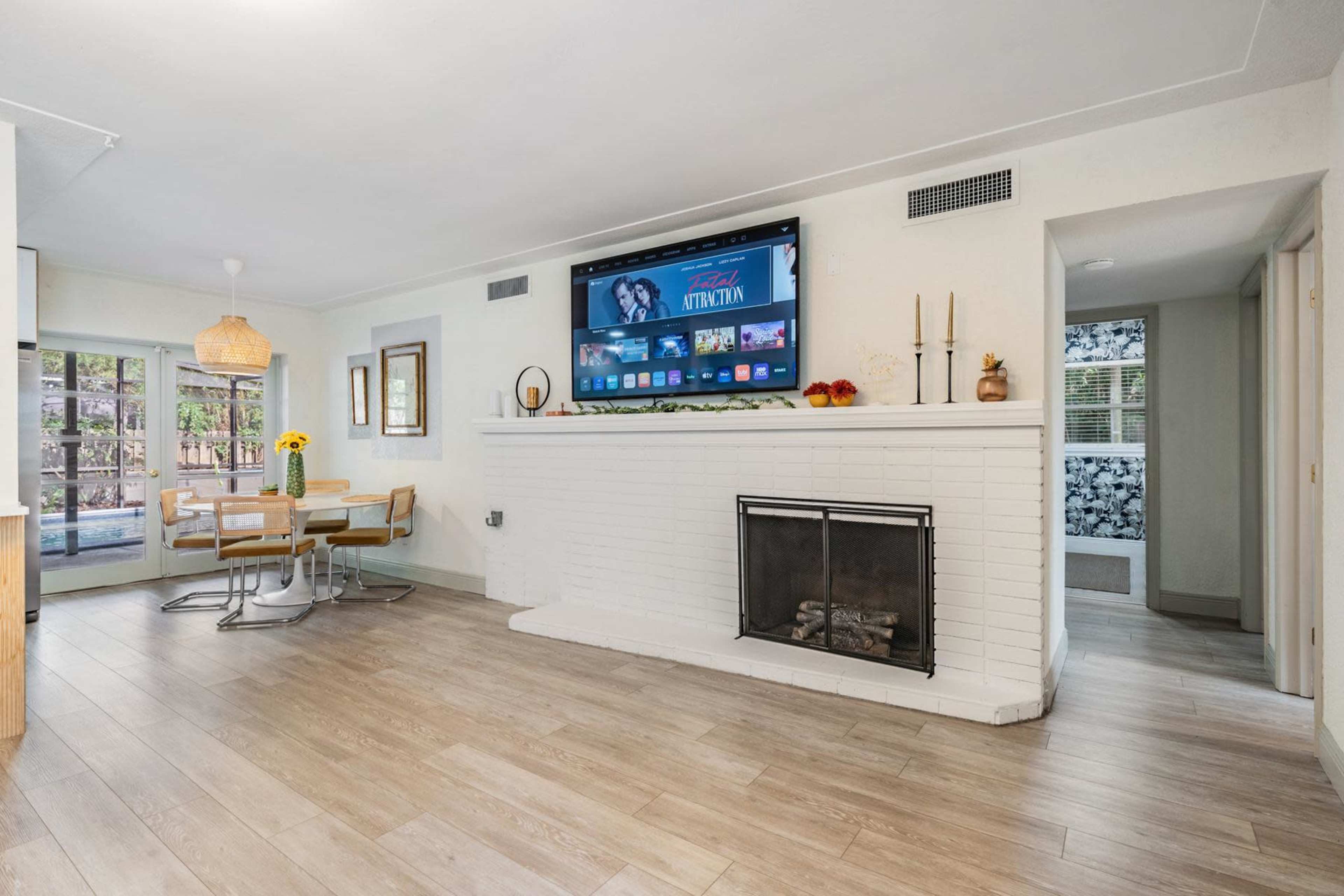 A living area featuring a fireplace, a large TV mounted on a white brick wall, and a dining table with chairs near a window.