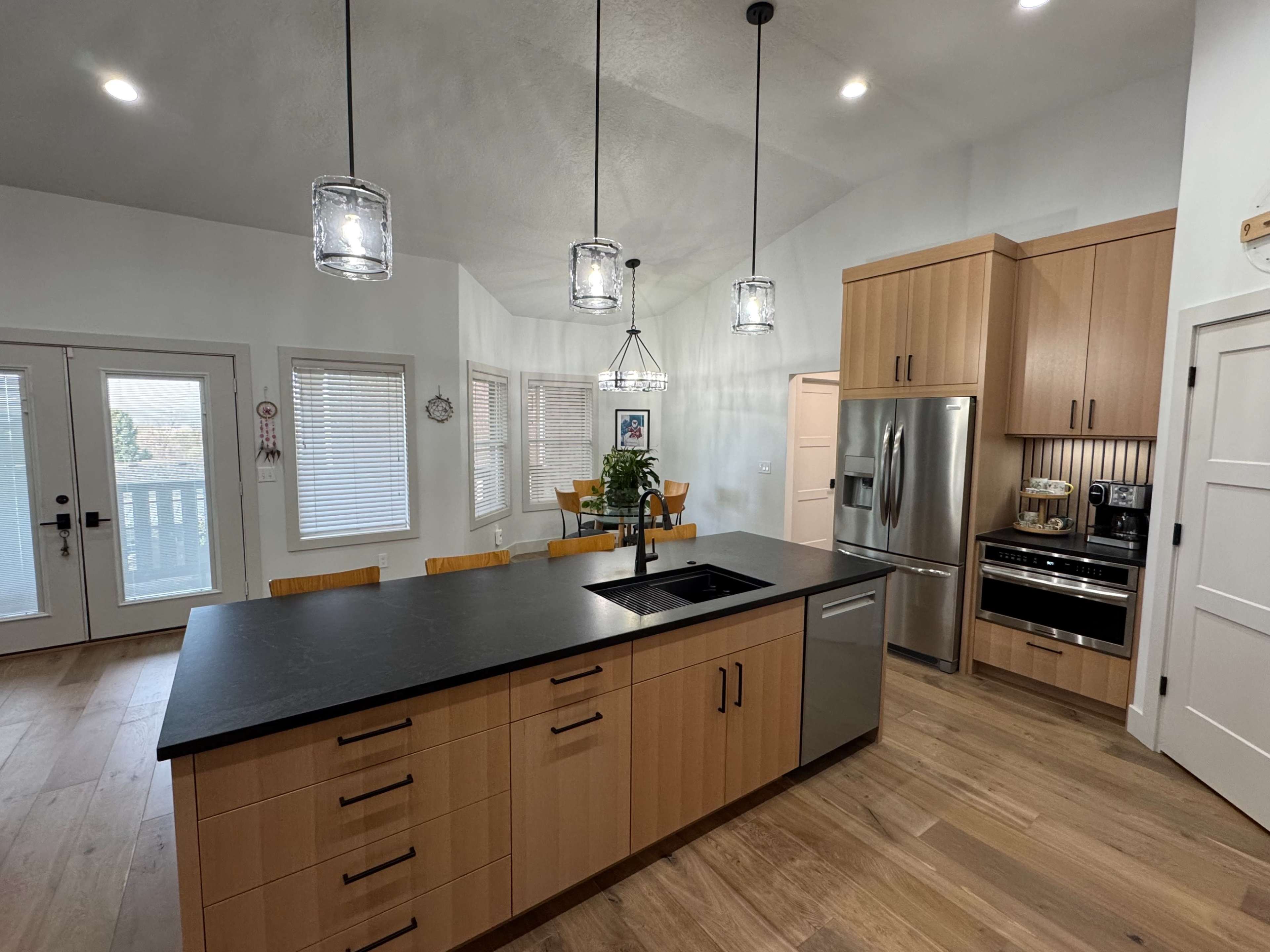 The image shows a modern kitchen with wooden cabinetry, a large black countertop island, stainless steel appliances, and a dining area visible in the background.