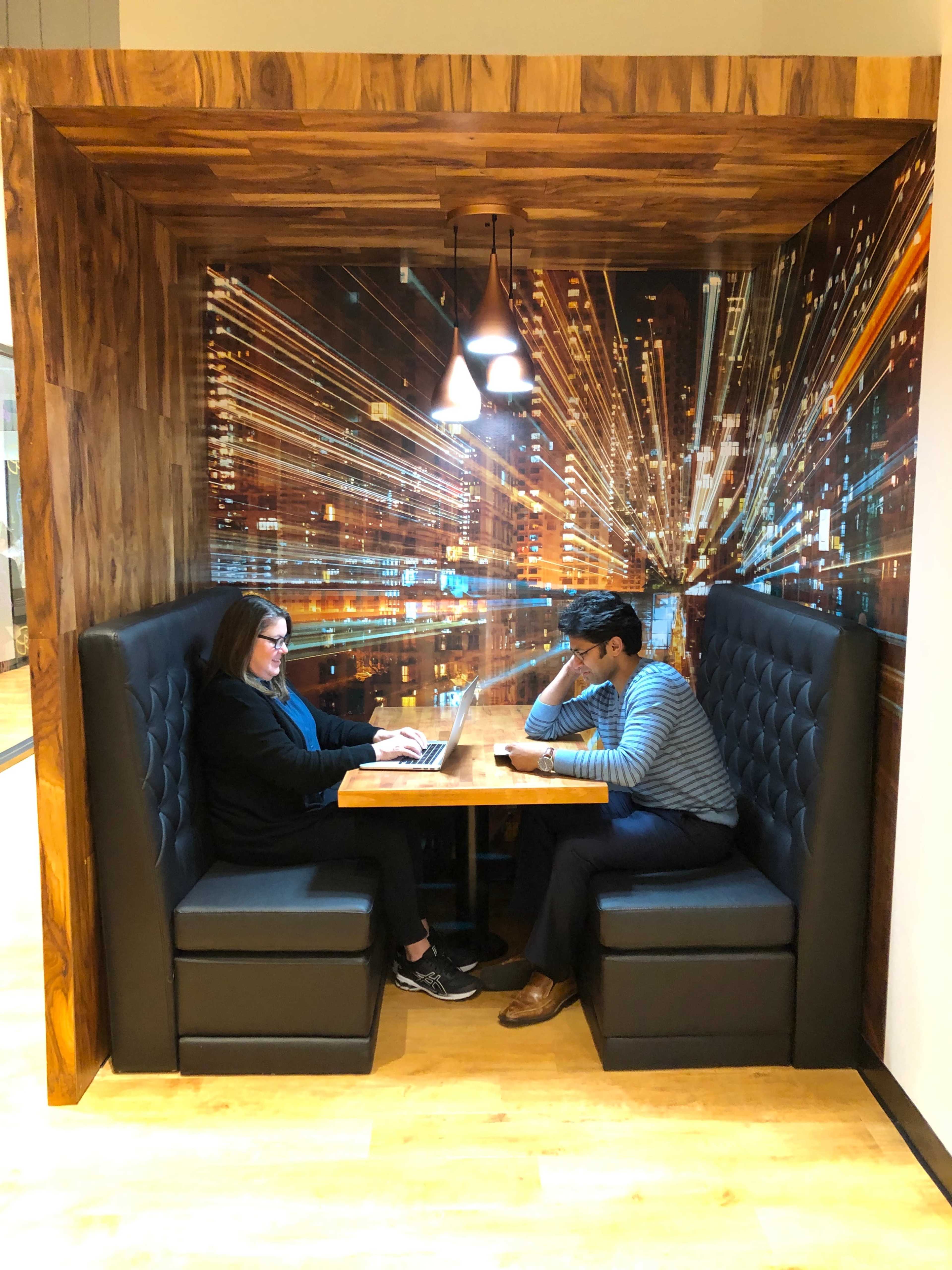 Two individuals are seated at a wooden table in a booth, one working on a laptop while the other appears to be in contemplation, with a cityscape mural in the background.