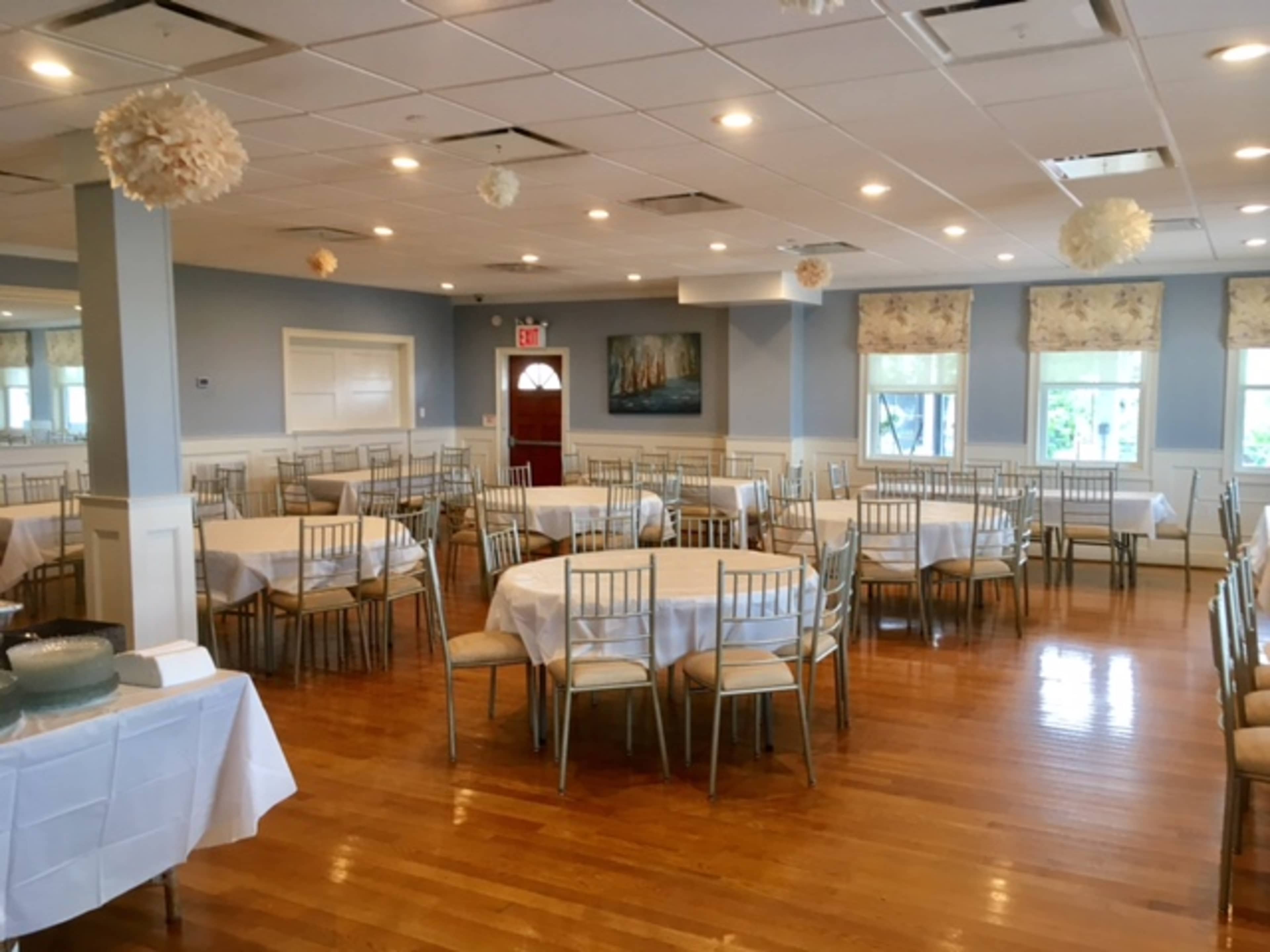 A spacious banquet hall features several round tables covered in white linens, surrounded by wooden chairs, with decorative paper flowers hanging from the ceiling.