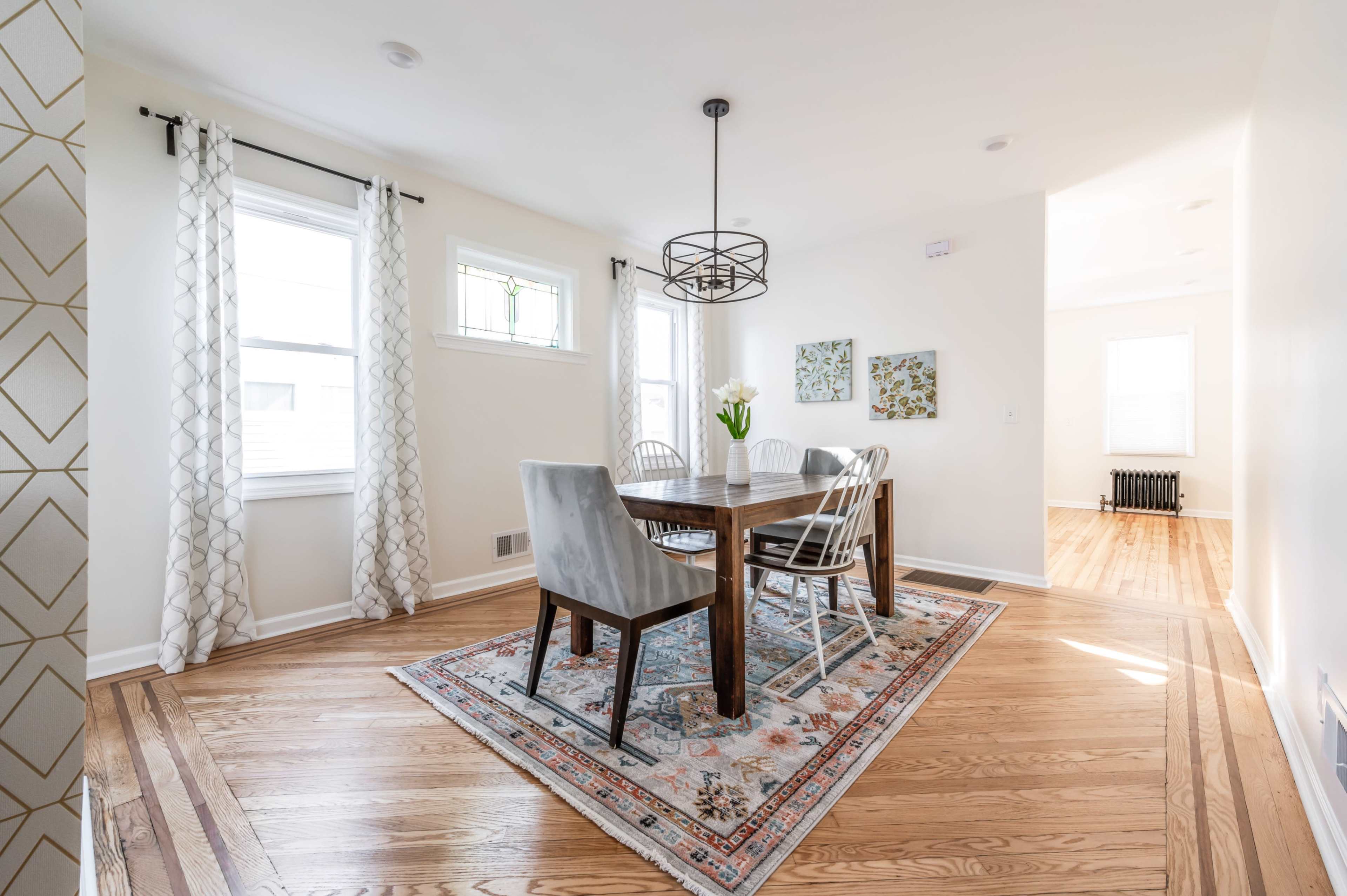 A dining area features a round wooden table surrounded by four chairs, a vase of flowers, and a decorative area rug beneath it, with windows allowing natural light to enter.
