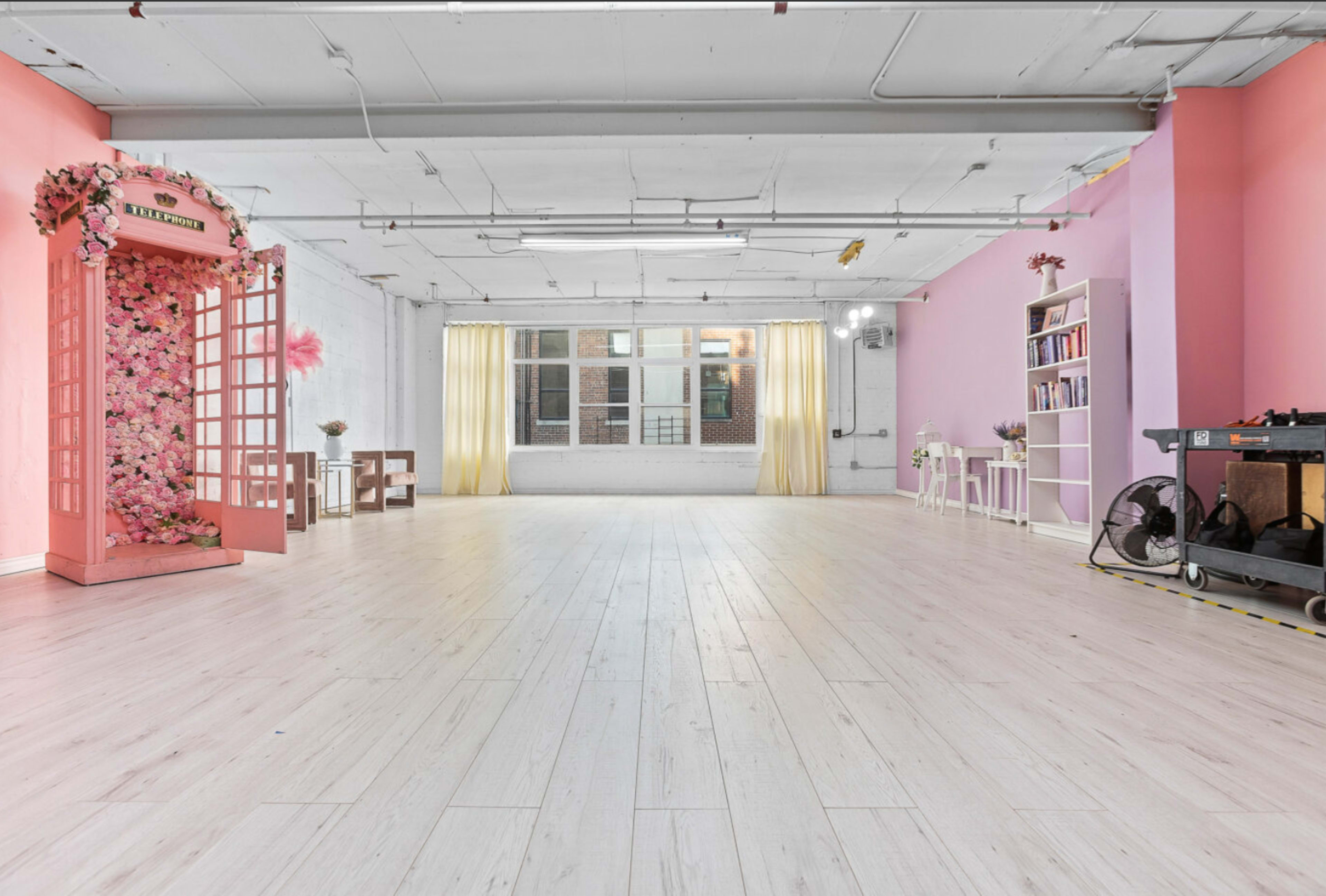 A spacious, brightly lit room with pink and white walls, featuring a floral-decorated entrance, large windows, and simple furniture.