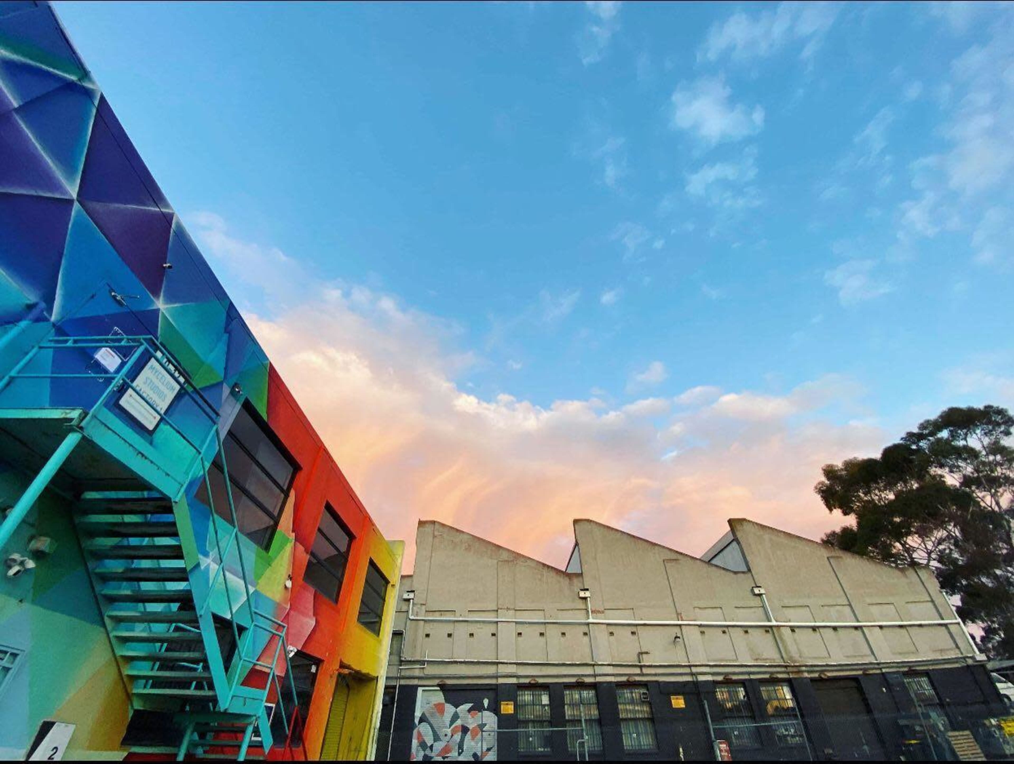 A vibrant mural on a building with colorful geometric patterns, alongside a gray industrial structure under a partly cloudy sky.