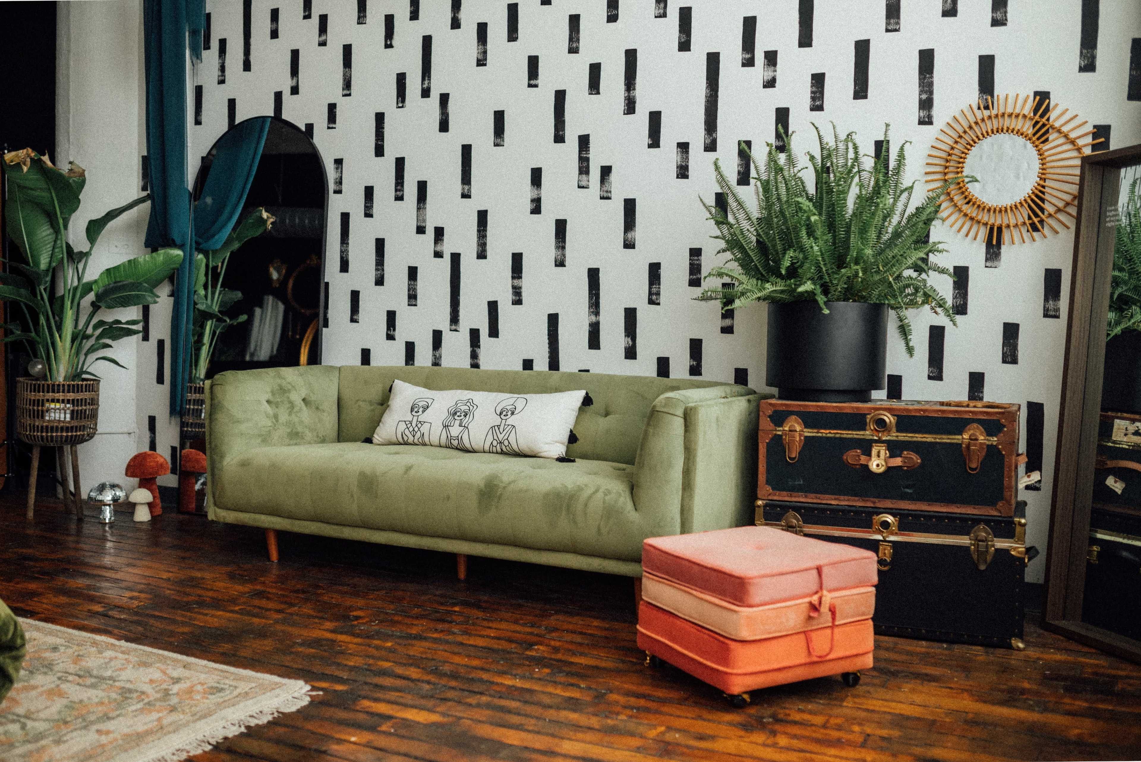 A green sofa with a patterned wall behind it, accompanied by a decorative mirror, a potted plant, and stacked vintage trunks.