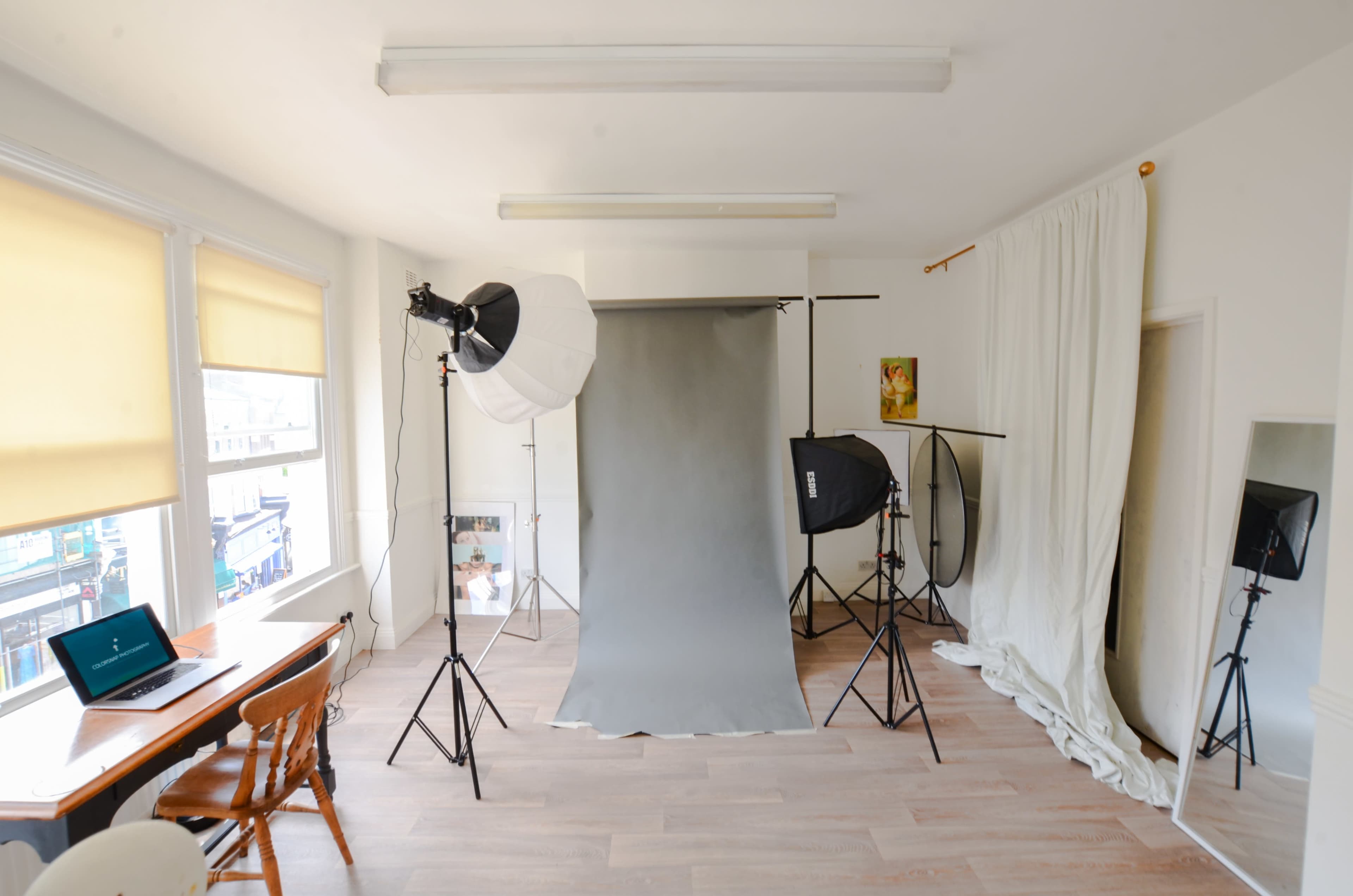 The image shows a well-lit photography studio with a gray backdrop, various lighting equipment, and a wooden table with a laptop.