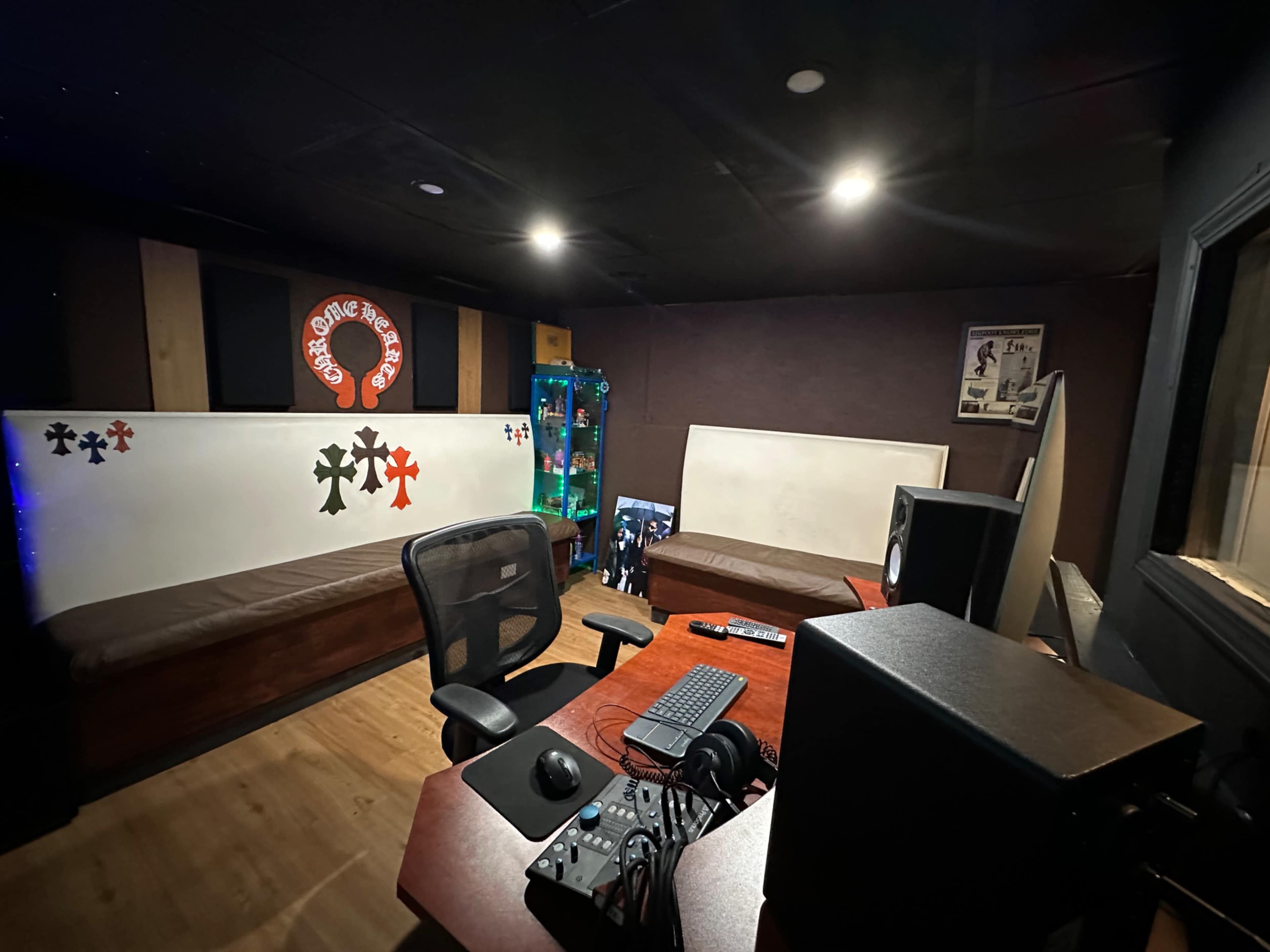 The image shows a small recording studio with a wooden desk, a computer monitor, audio equipment, and a seating area on either side.