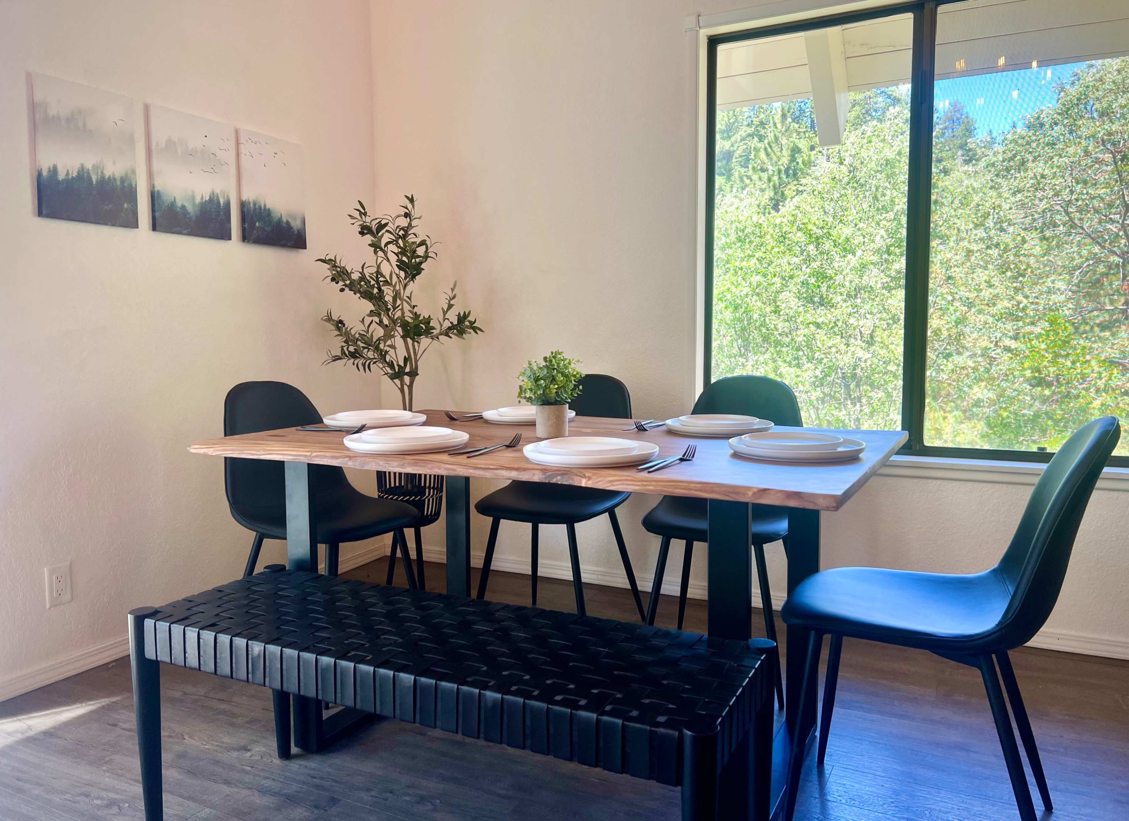 A dining area features a wooden table set with plates, surrounded by black chairs and a bench, with greenery visible through a large window.