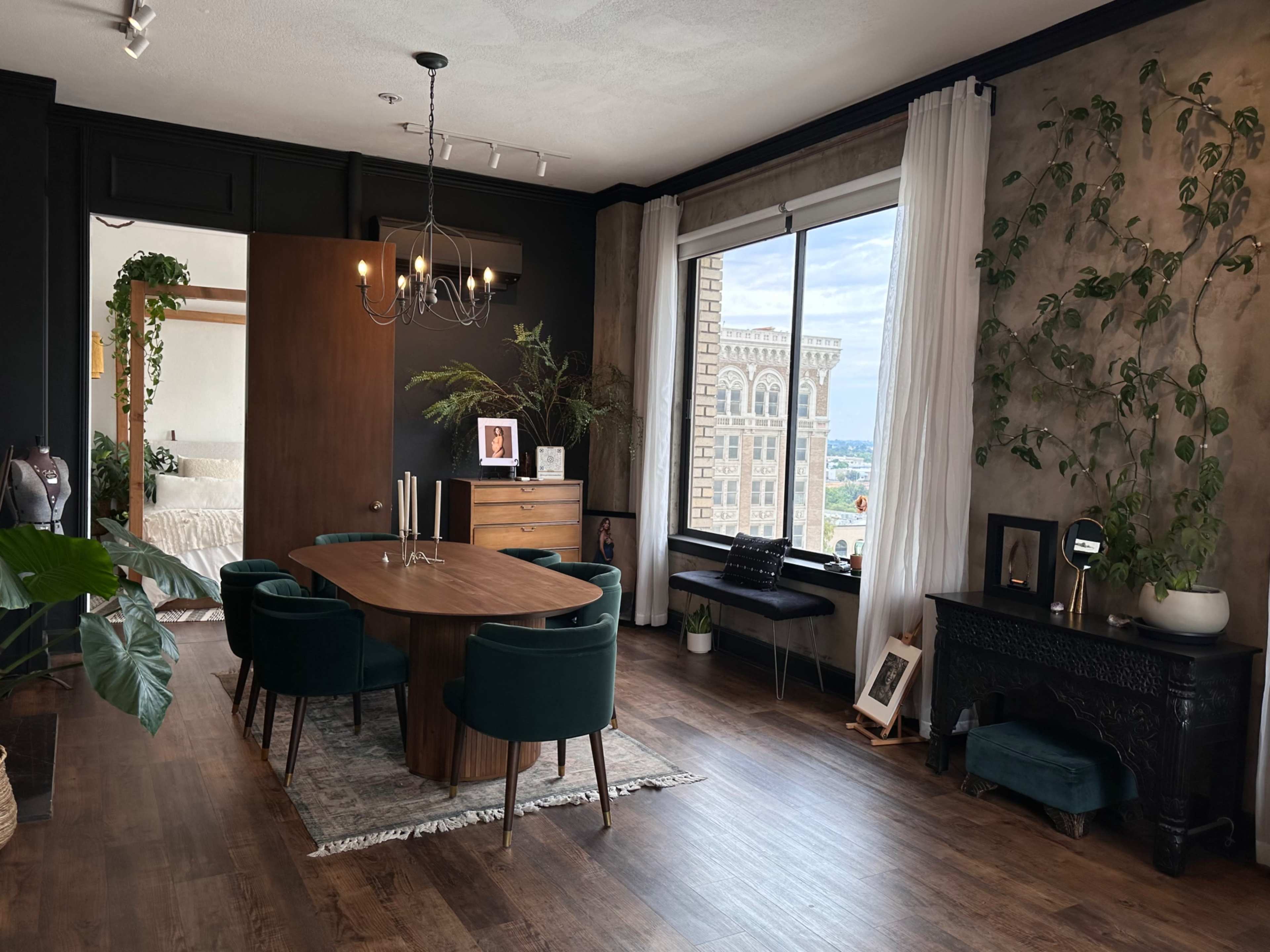 A modern dining area with a round table, green upholstered chairs, a wood dresser, and a large window overlooking a cityscape.