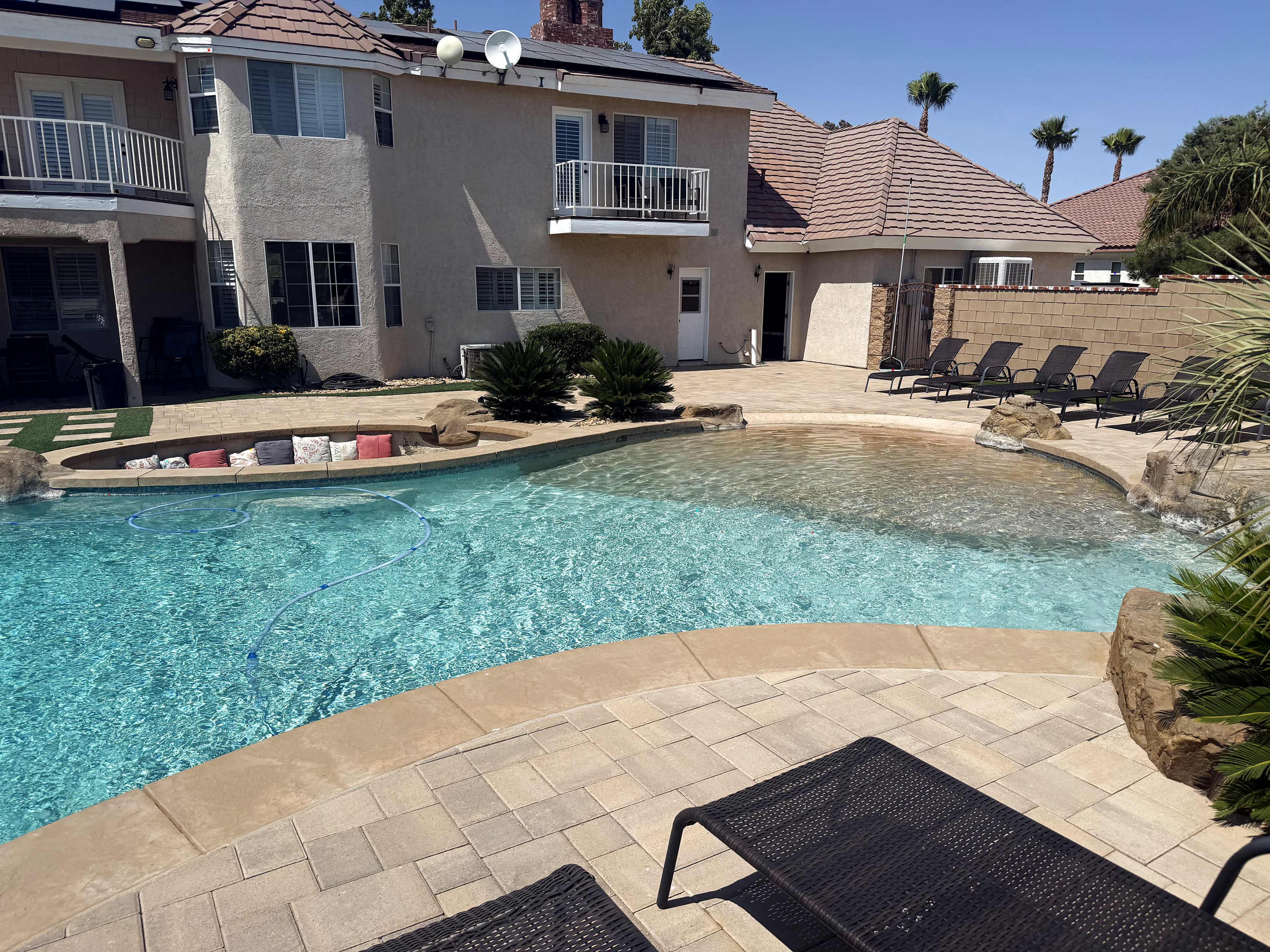 The image shows a backyard with a pool, two lounge chairs, and a two-story house with balconies under a clear blue sky.