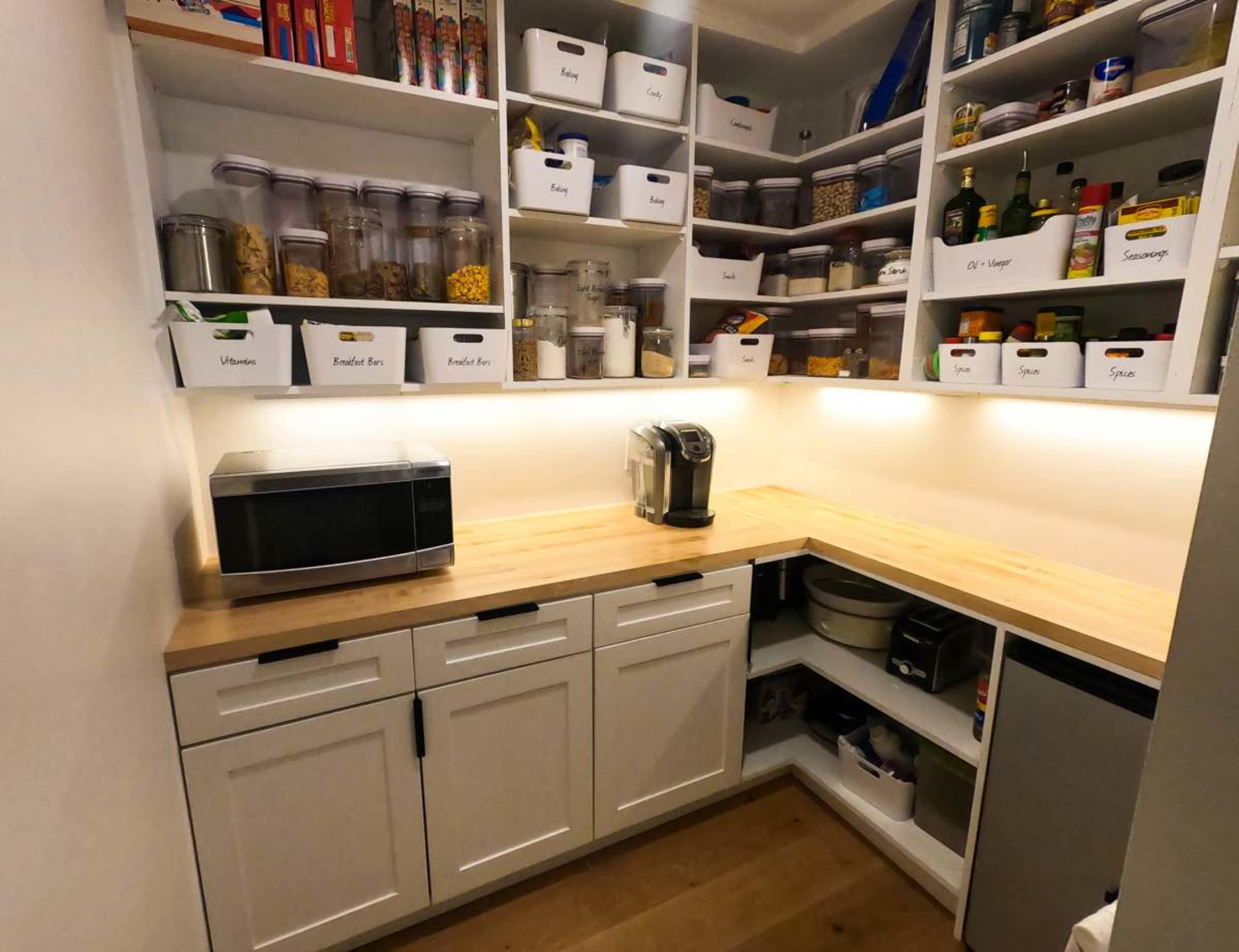 A well-organized pantry features shelves filled with labeled containers of dry goods, spices, and a countertop housing a microwave and coffee maker.