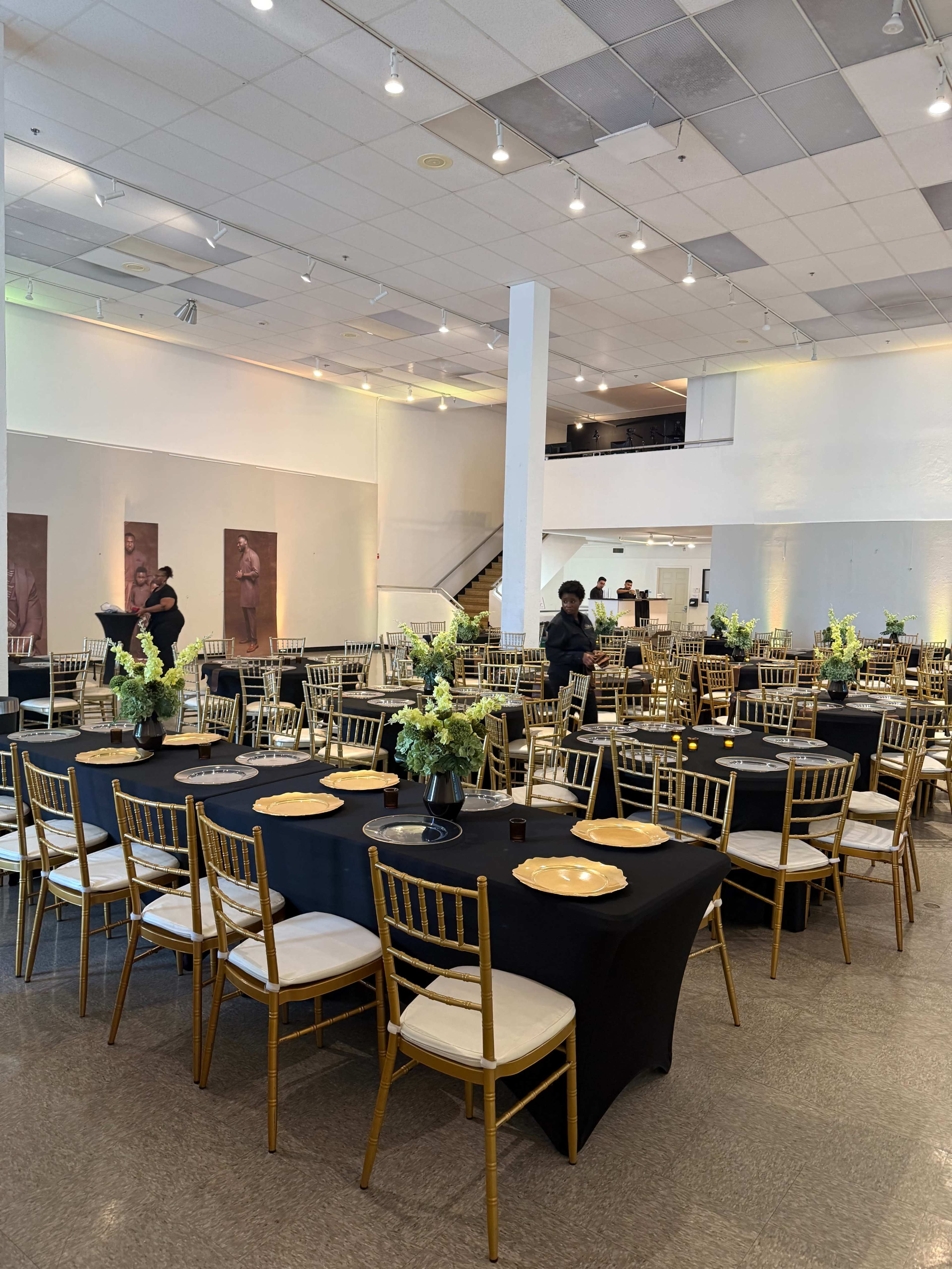 A spacious event hall is set up with black tablecloths and gold chairs, featuring floral centerpieces on each table and a staff member arranging the seating.