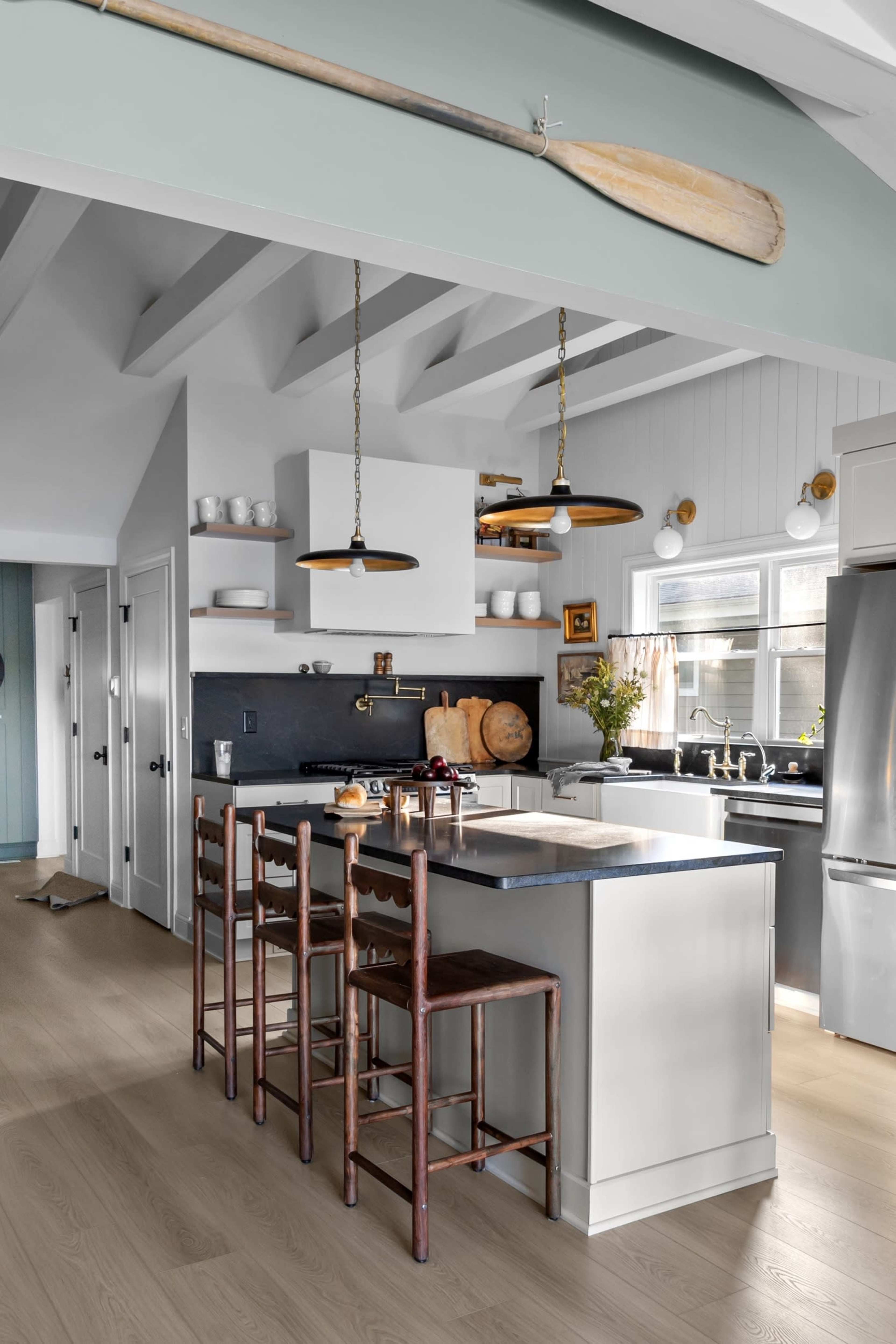 The image shows a modern kitchen with a central island featuring wooden stools, hanging pendant lights, and white cabinetry, all under a ceiling with exposed beams.