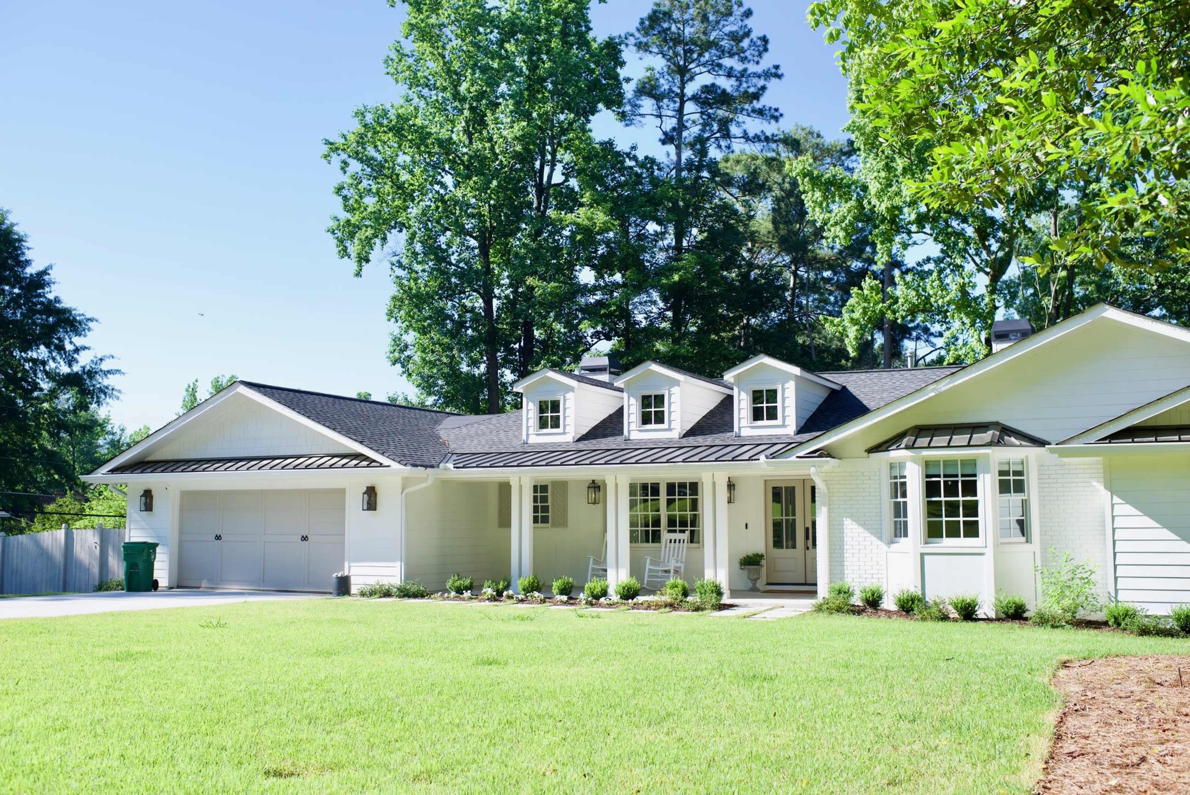 A single-story white house with a gray roof and three dormer windows is set in a grassy yard surrounded by tall trees.