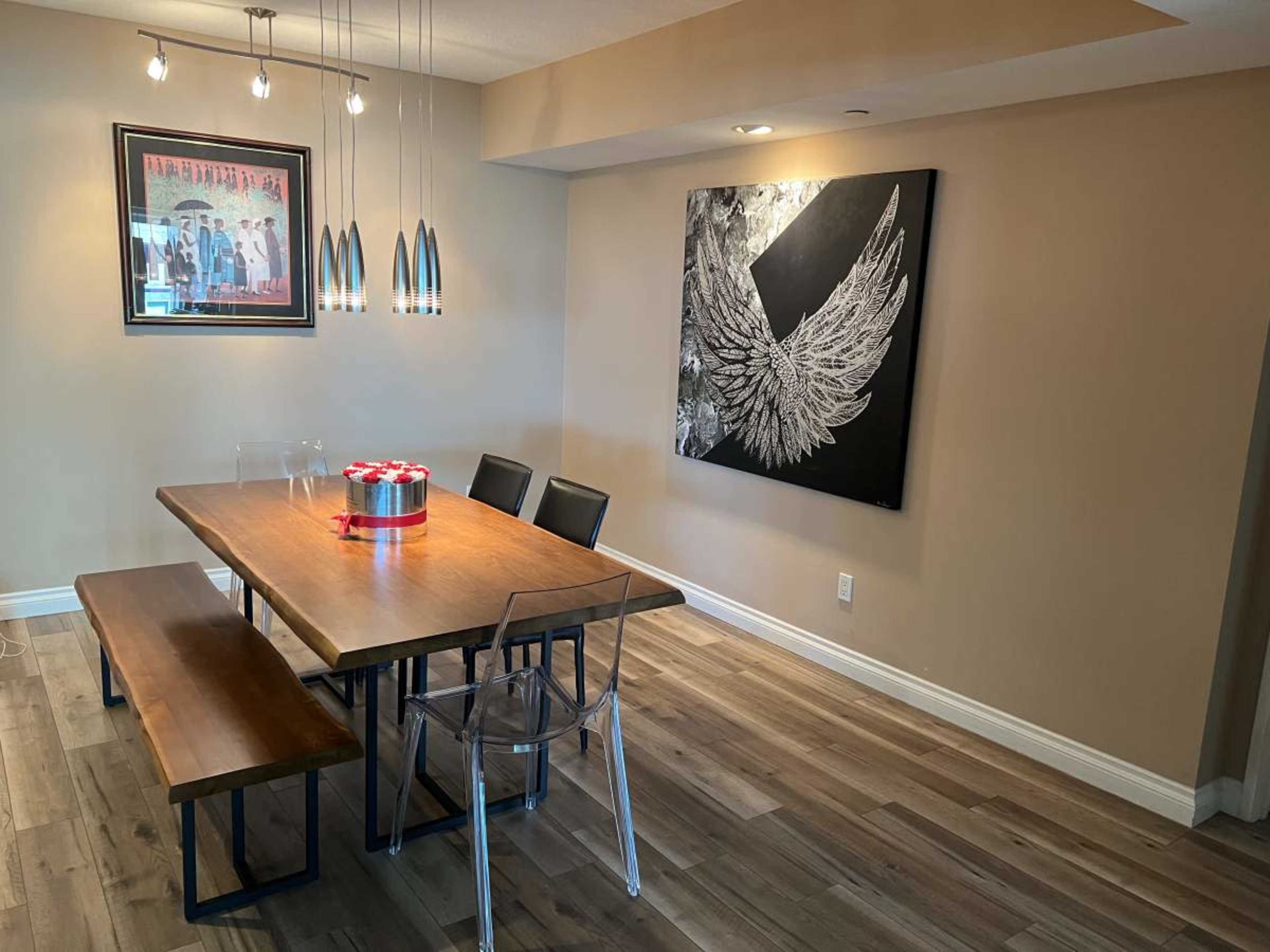 A dining area with a wooden table, clear acrylic chairs, a bench, and a large winged artwork on the wall.