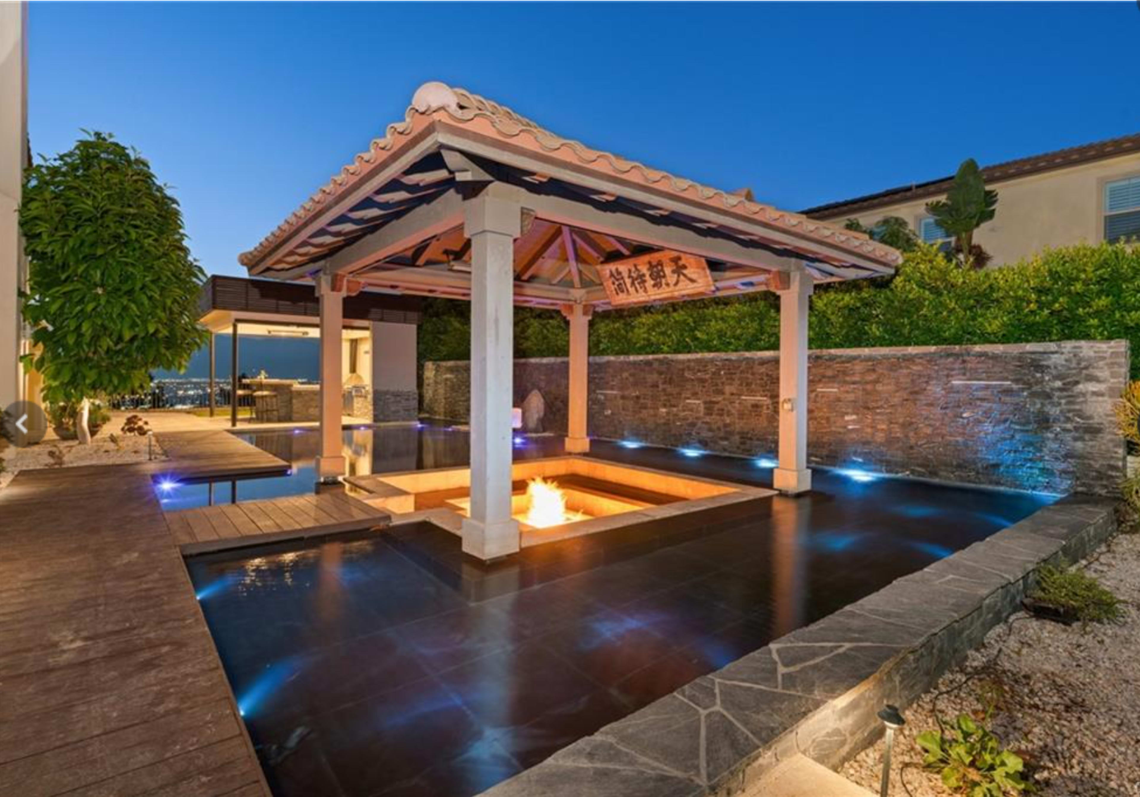 The image features a modern outdoor space with a traditional-style pavilion over a square hot tub, surrounded by stone walls and illuminated pathways.