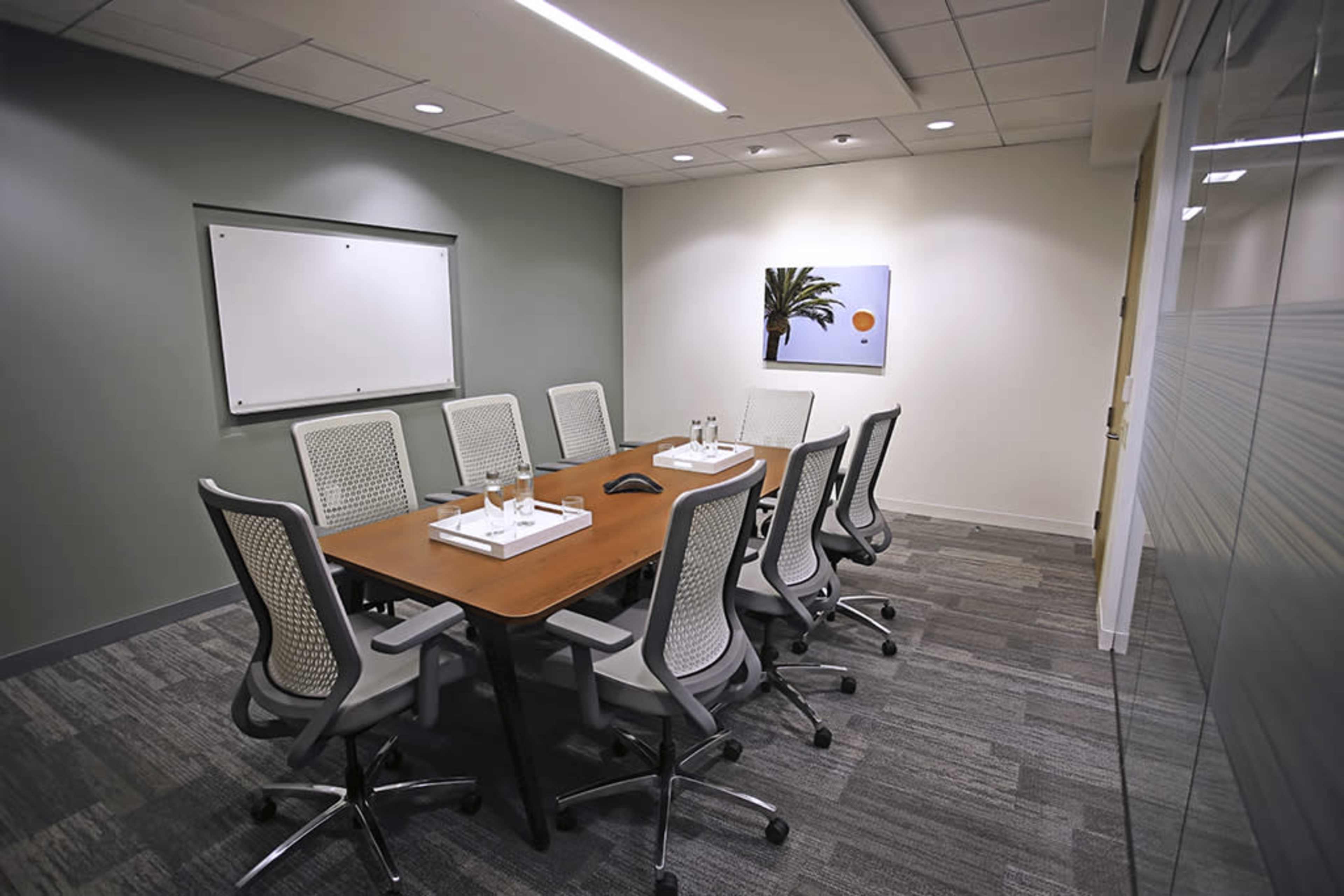 A conference room features a long wooden table surrounded by ten ergonomic chairs, with a large whiteboard and an abstract painting on the wall.