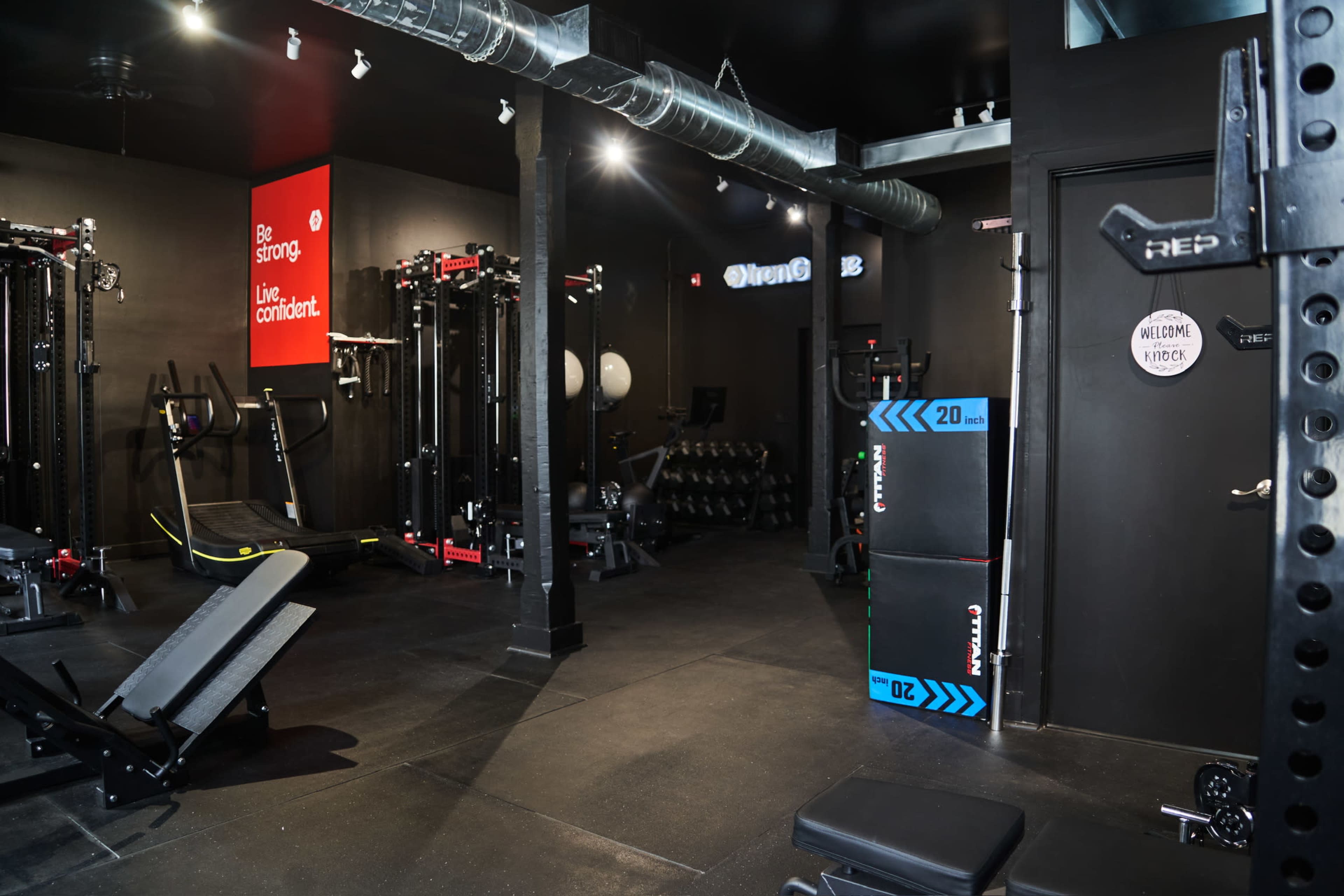 The image shows a modern gym interior with various fitness equipment, including weights, benches, and exercise machines, set against black walls.