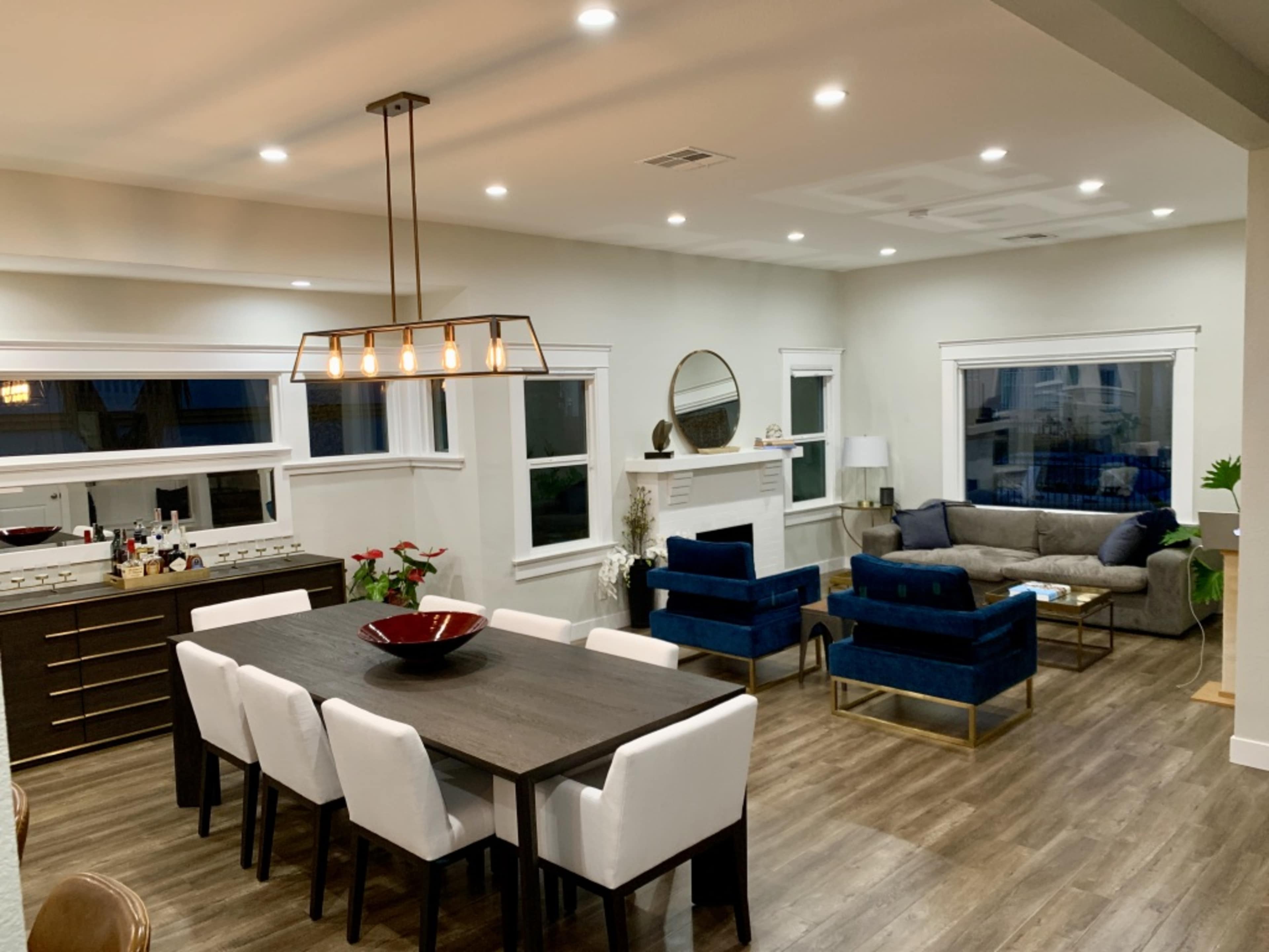 The image shows a modern living and dining area with a large table and chairs, a sofa, blue armchairs, and a fireplace in a well-lit space.