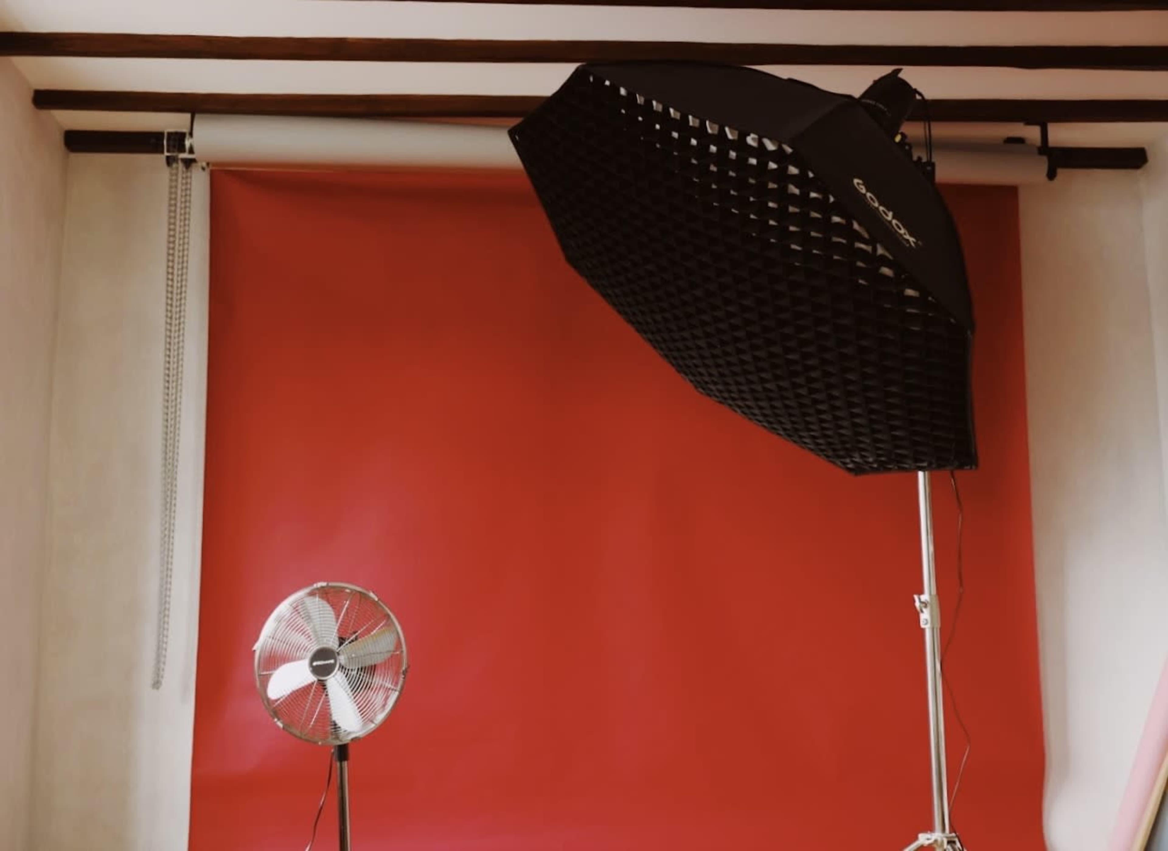 A photography studio is set up with a red backdrop, a large softbox light, and a standing fan.