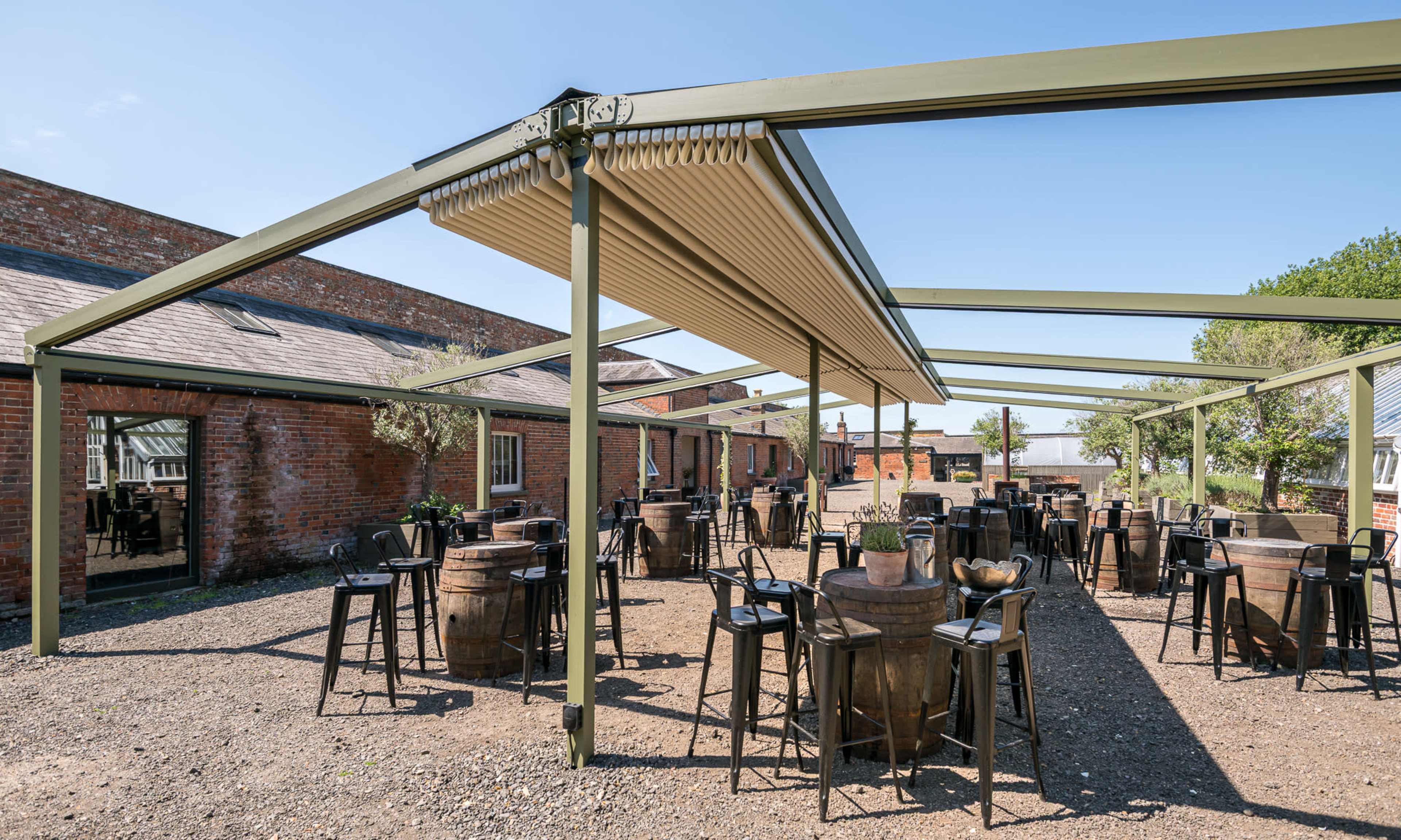 The image shows an outdoor seating area with tables and chairs arranged under a large canopy, surrounded by brick buildings and gravel ground.