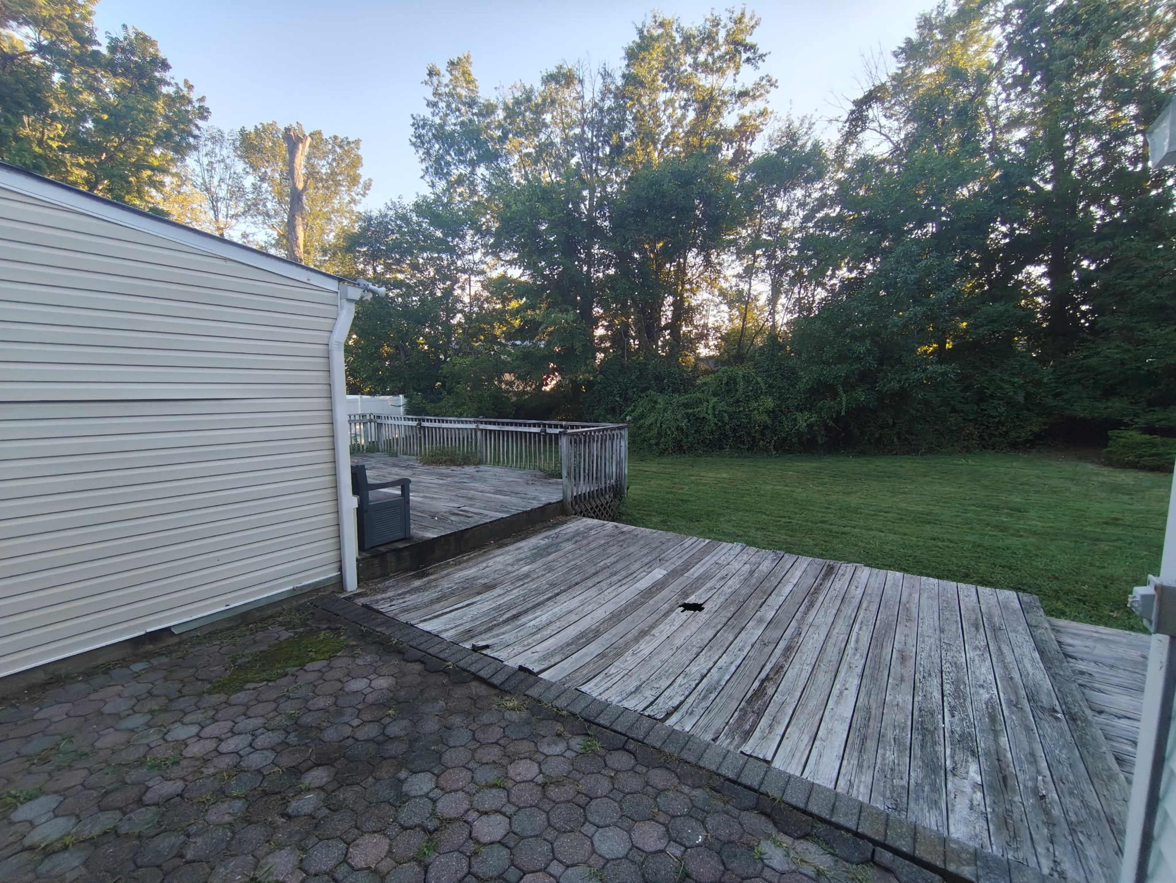 The image shows a wooden deck extending into a grassy area, surrounded by trees, with a paved section visible to the left.