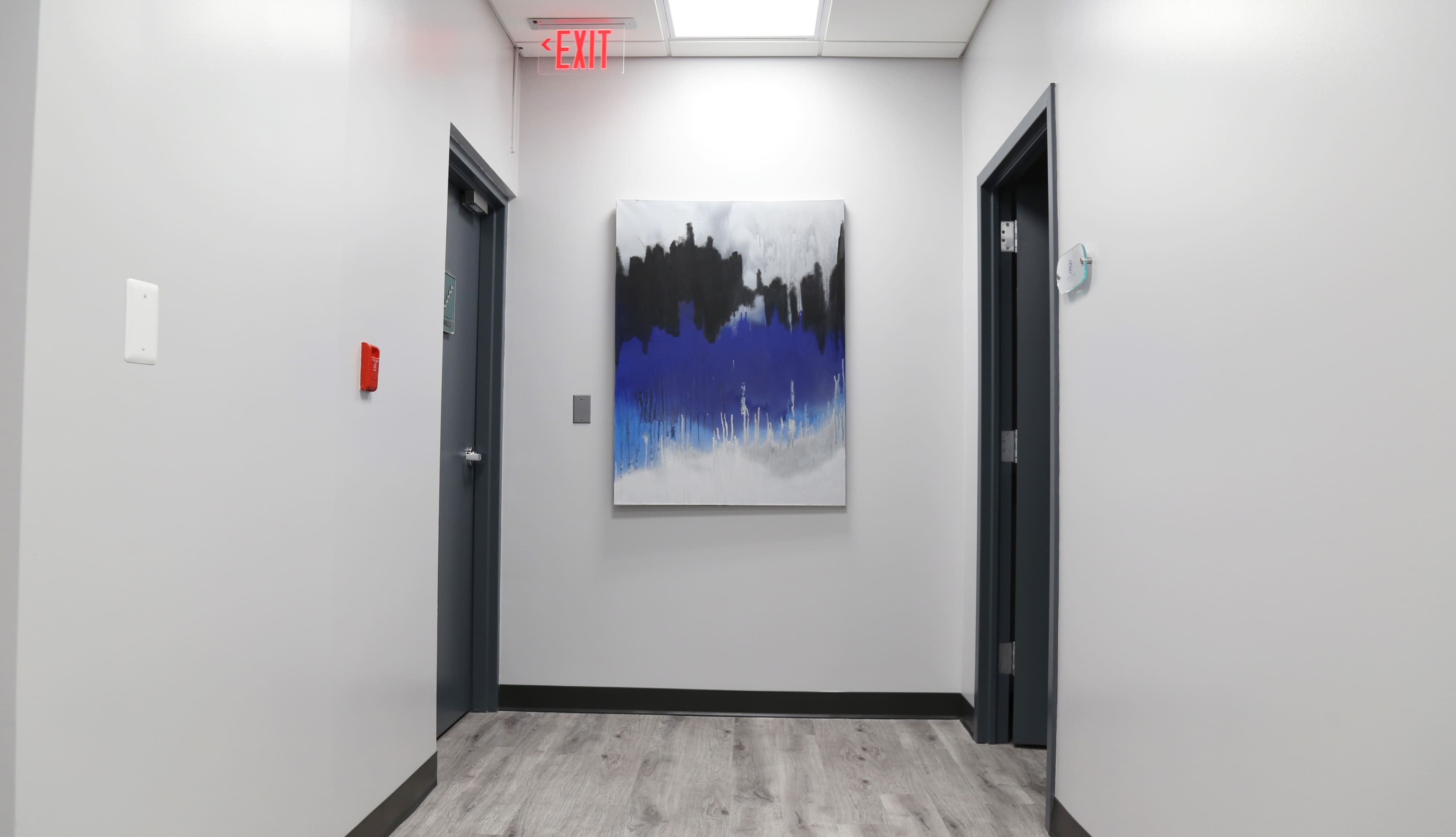 A hallway with grey walls, a wooden floor, and an abstract painting featuring blue, black, and white colors mounted on the wall between two doors.