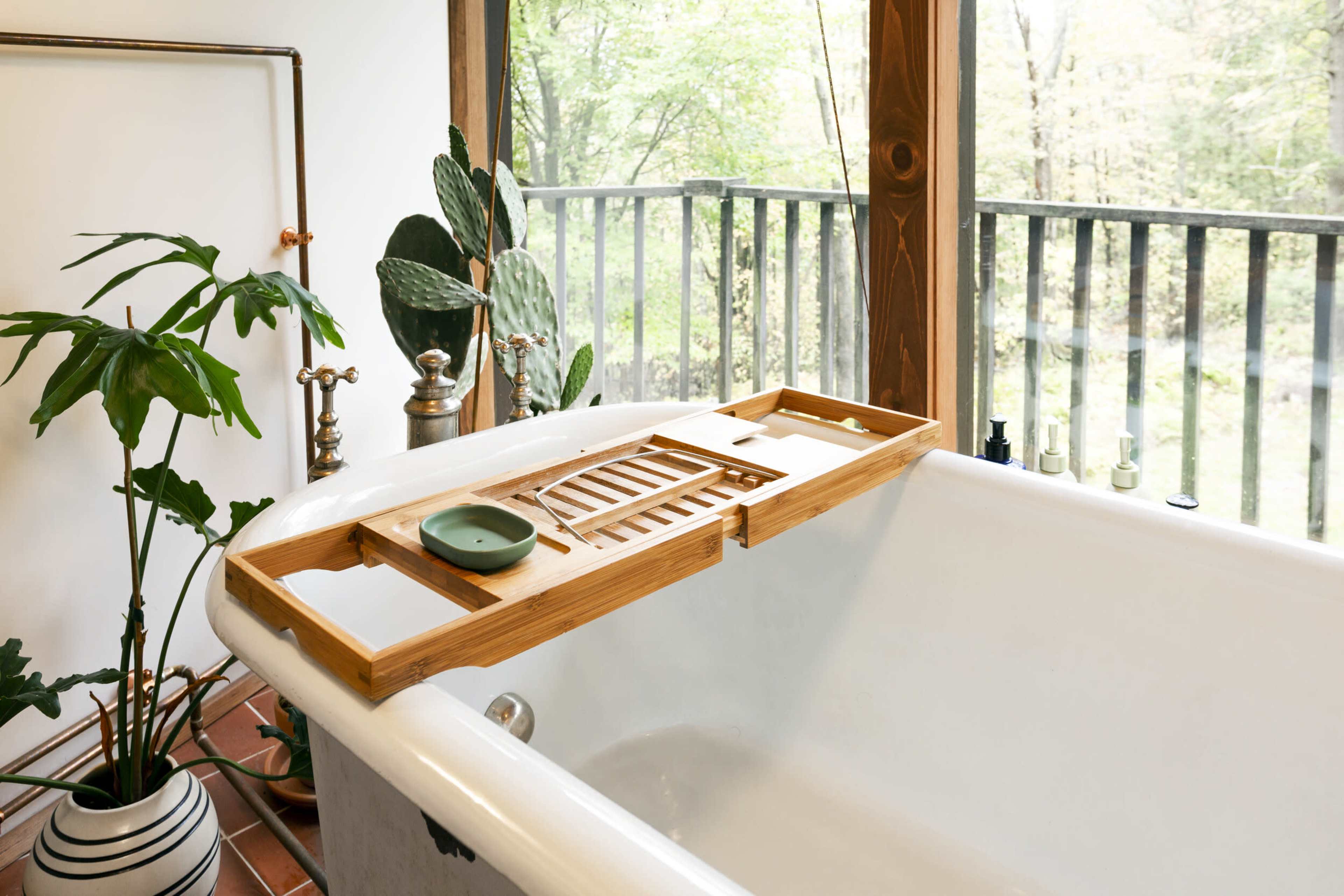 A wooden bathtub caddy rests on the edge of a white bathtub, surrounded by greenery and large windows showing a forest view.