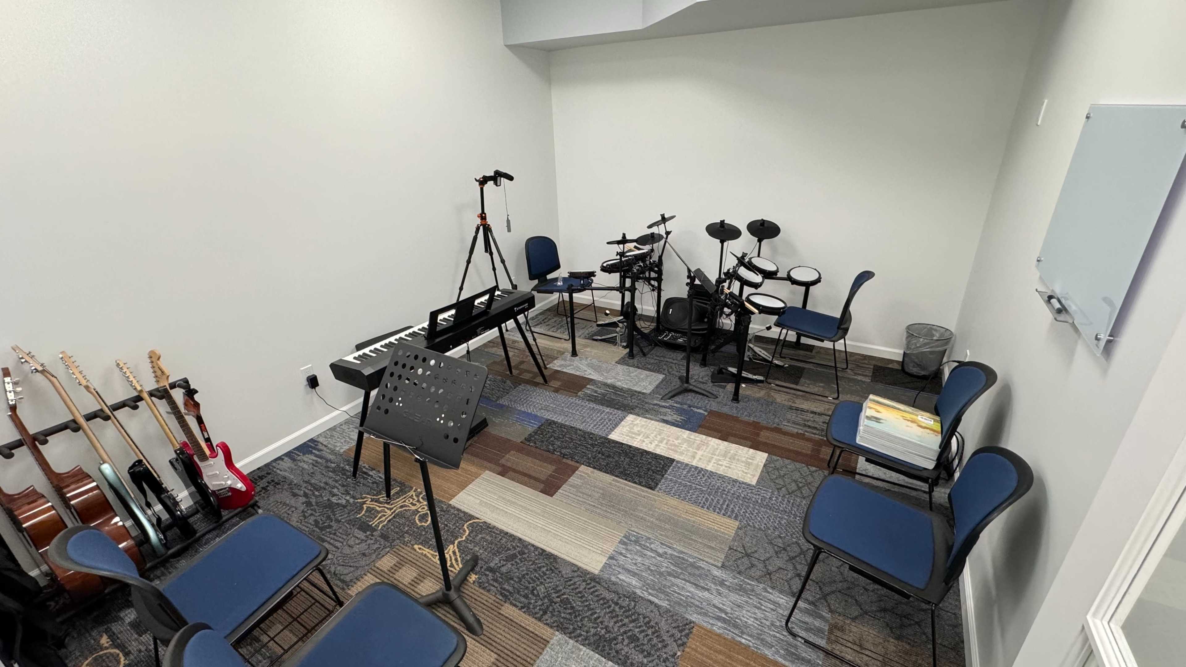 A small music room with a digital piano, electronic drum set, and several chairs arranged around a carpeted floor.