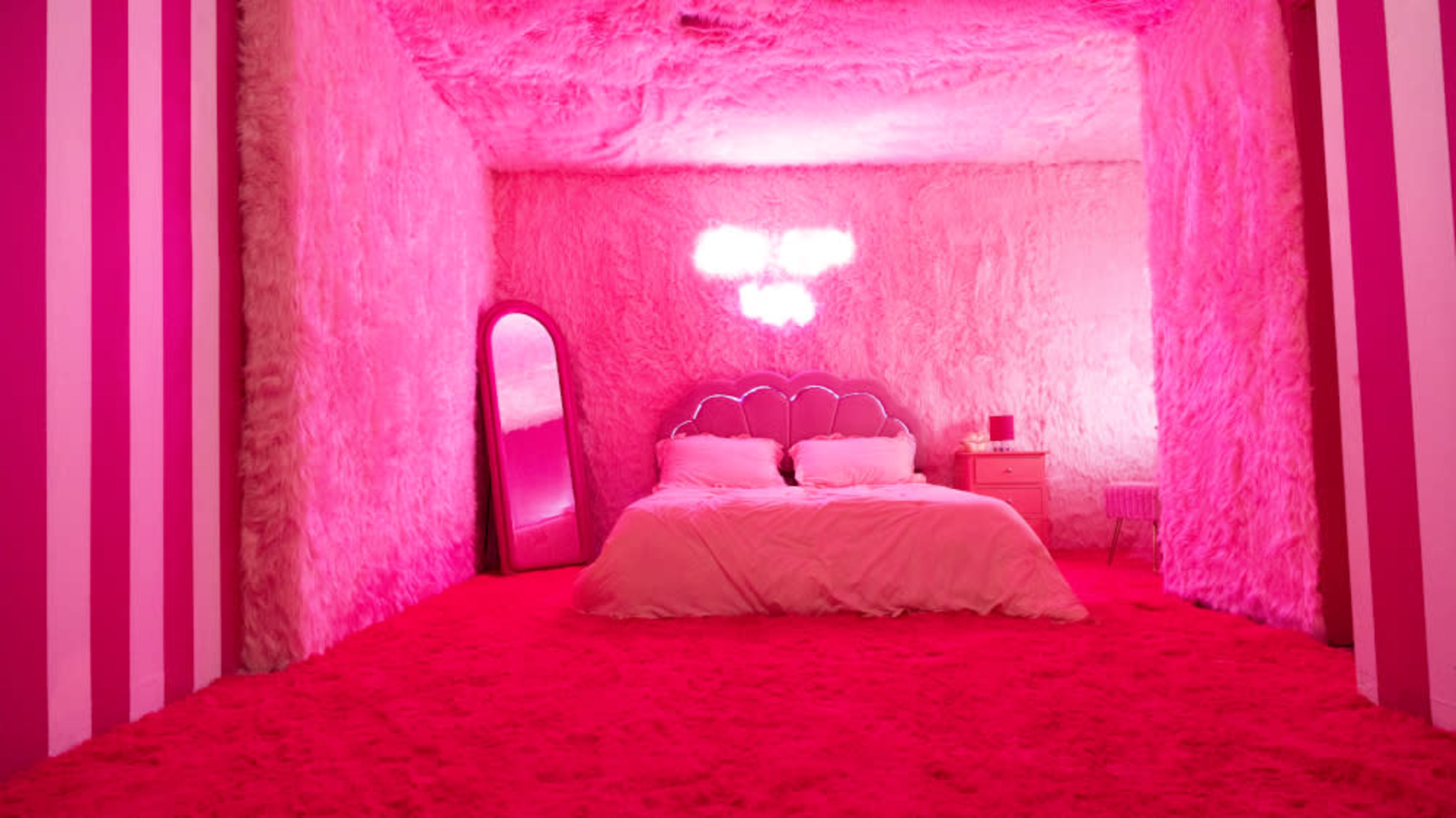 The image depicts a vibrant pink bedroom with fuzzy walls, a bed with a scalloped headboard, a round mirror, and a nightstand.