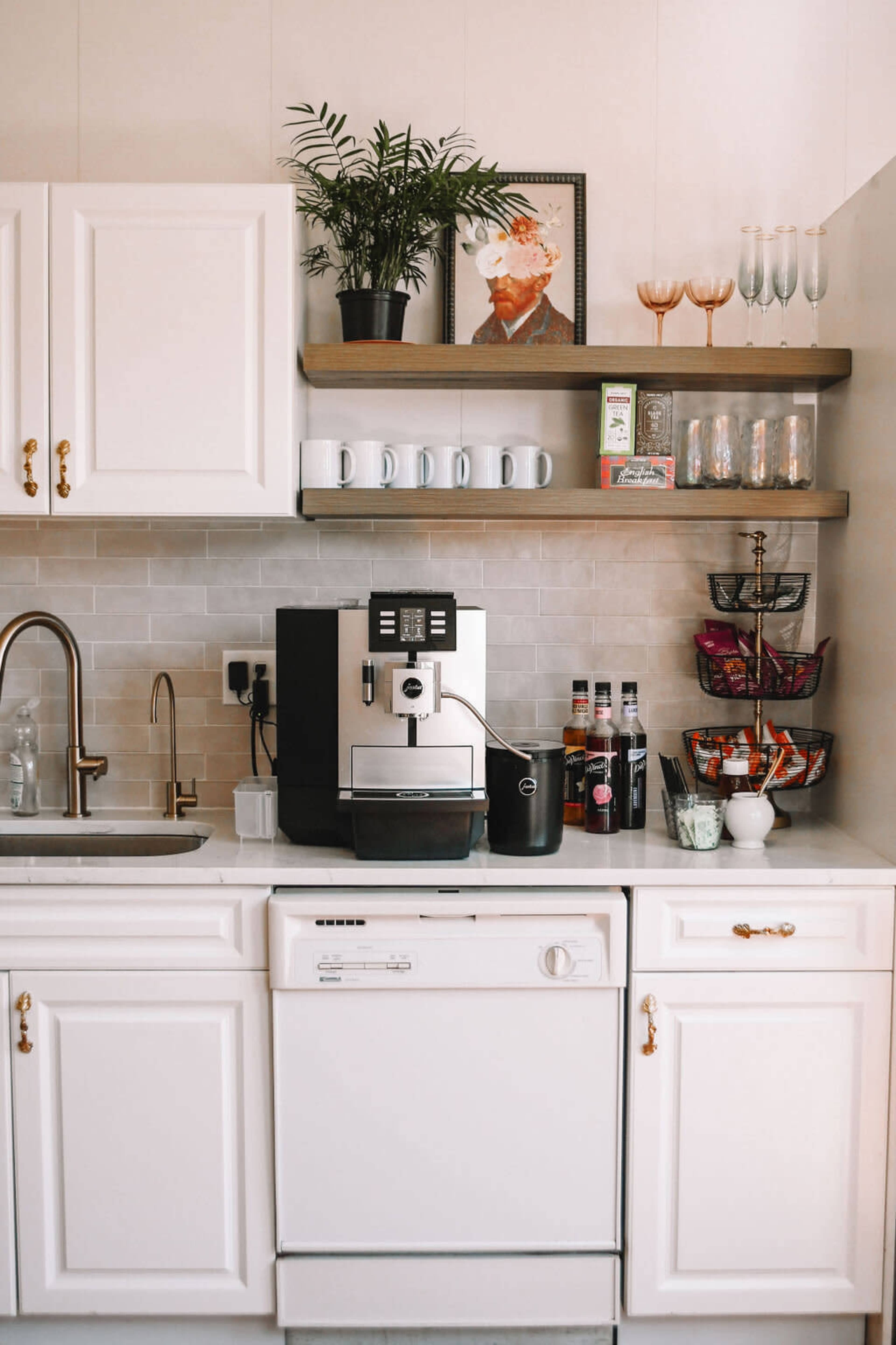 A modern kitchen setup features a sleek coffee machine, a set of white mugs, a dishwasher, and decorative items on open shelves.