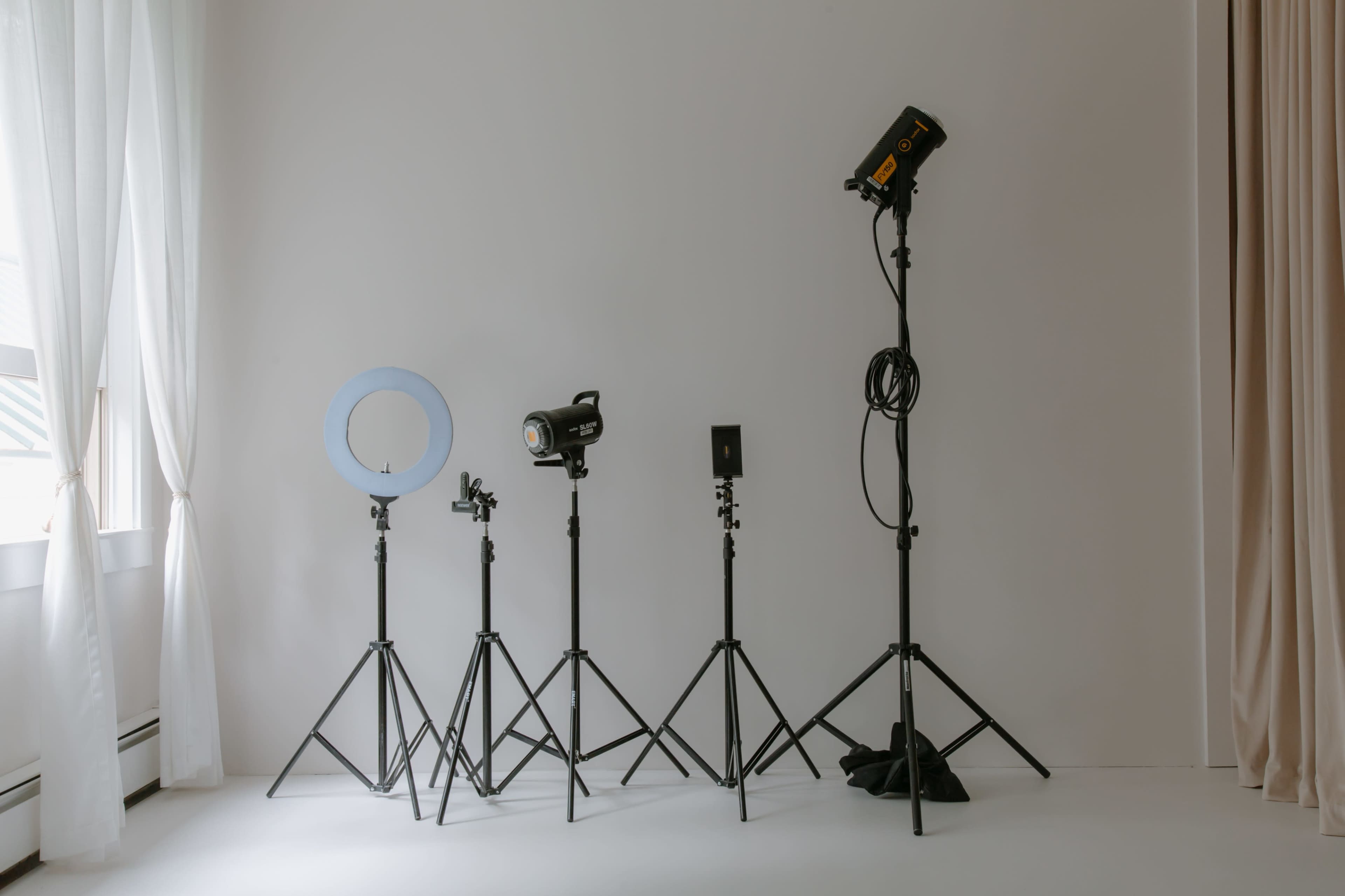 A row of various photography lights and equipment stands against a plain wall in a bright room.