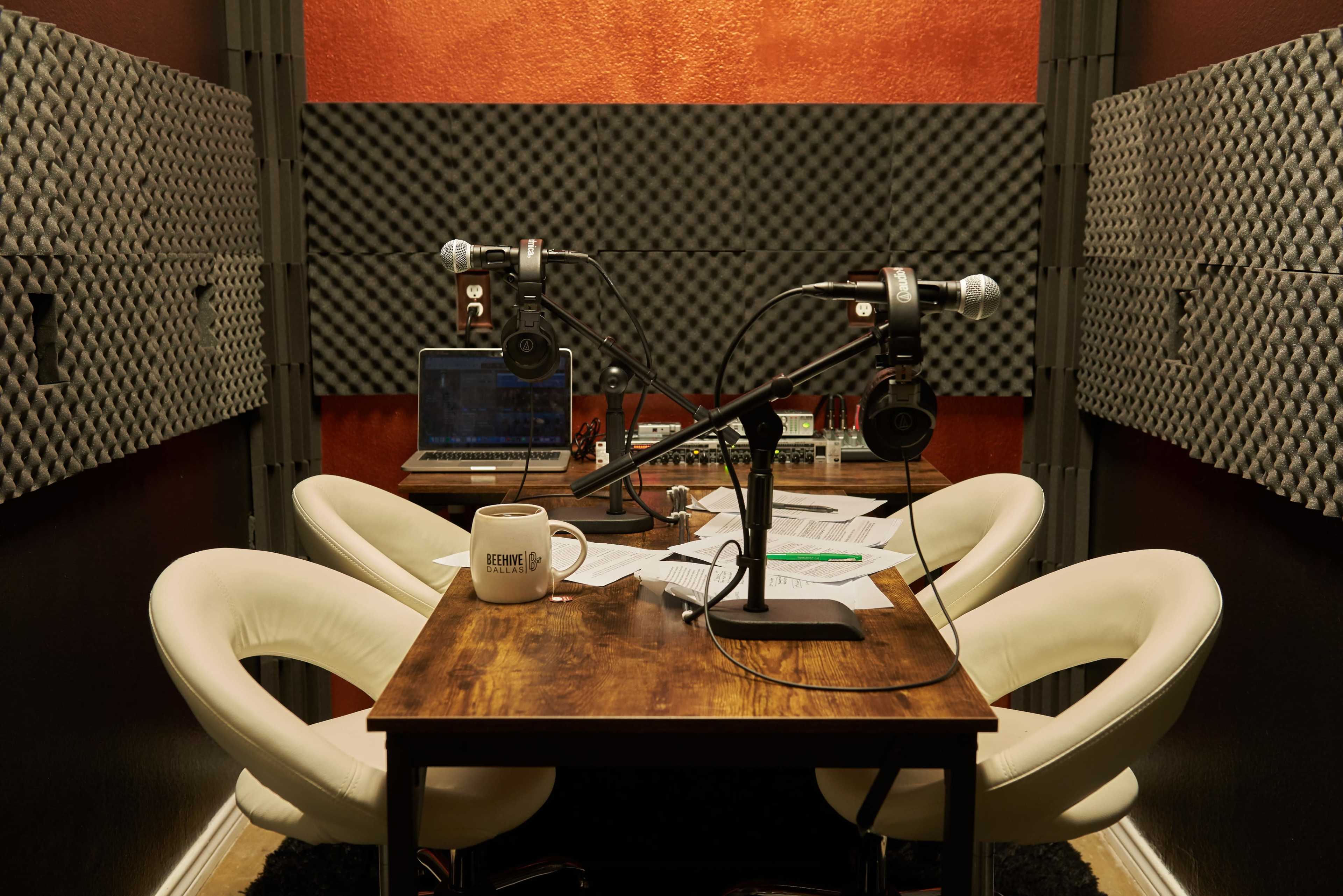 A small recording studio with two microphones, headphones, a laptop, and a table surrounded by acoustic foam panels.