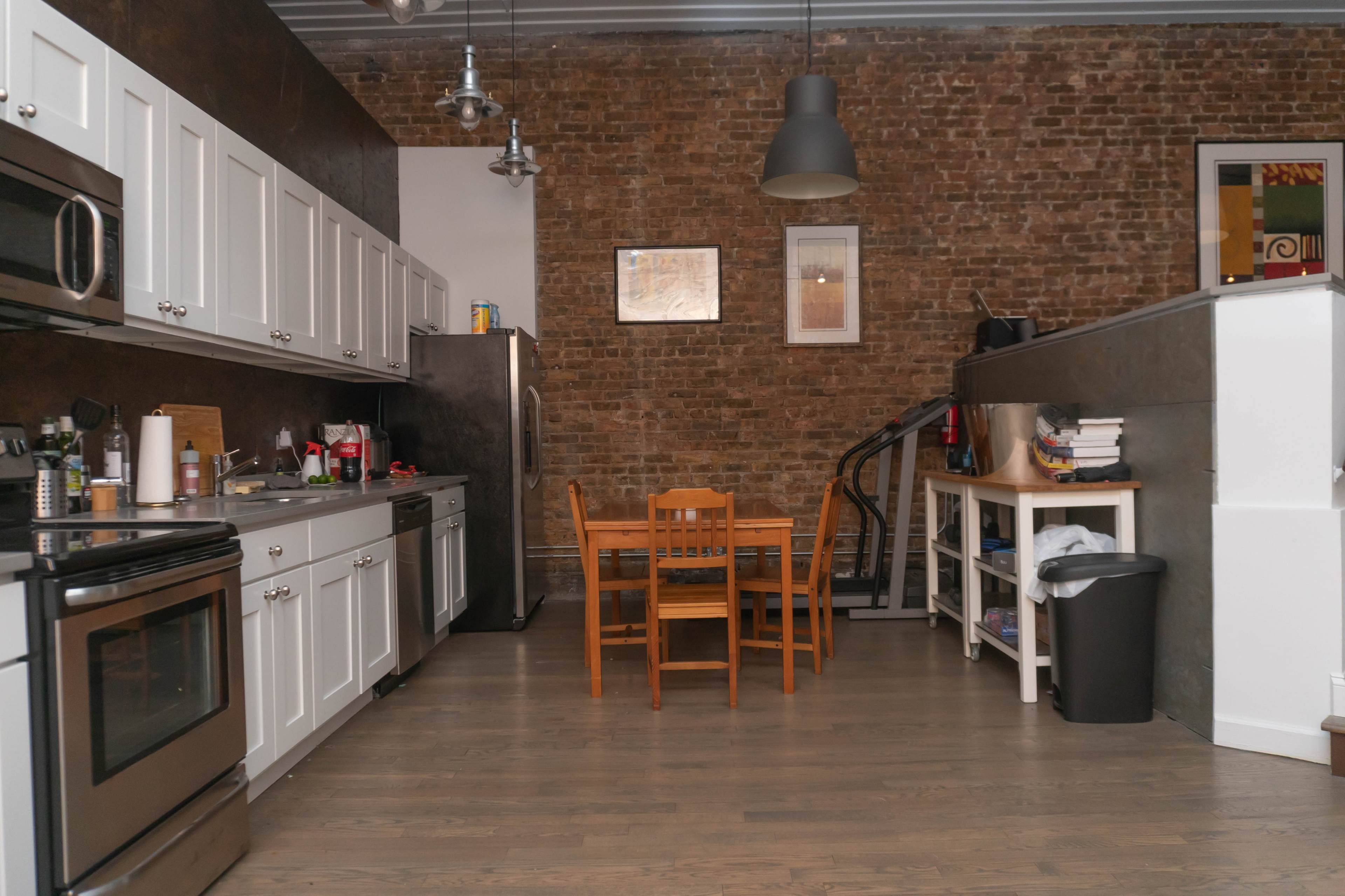 A modern kitchen features white cabinets, stainless steel appliances, a wooden dining table with chairs, and exposed brick walls.