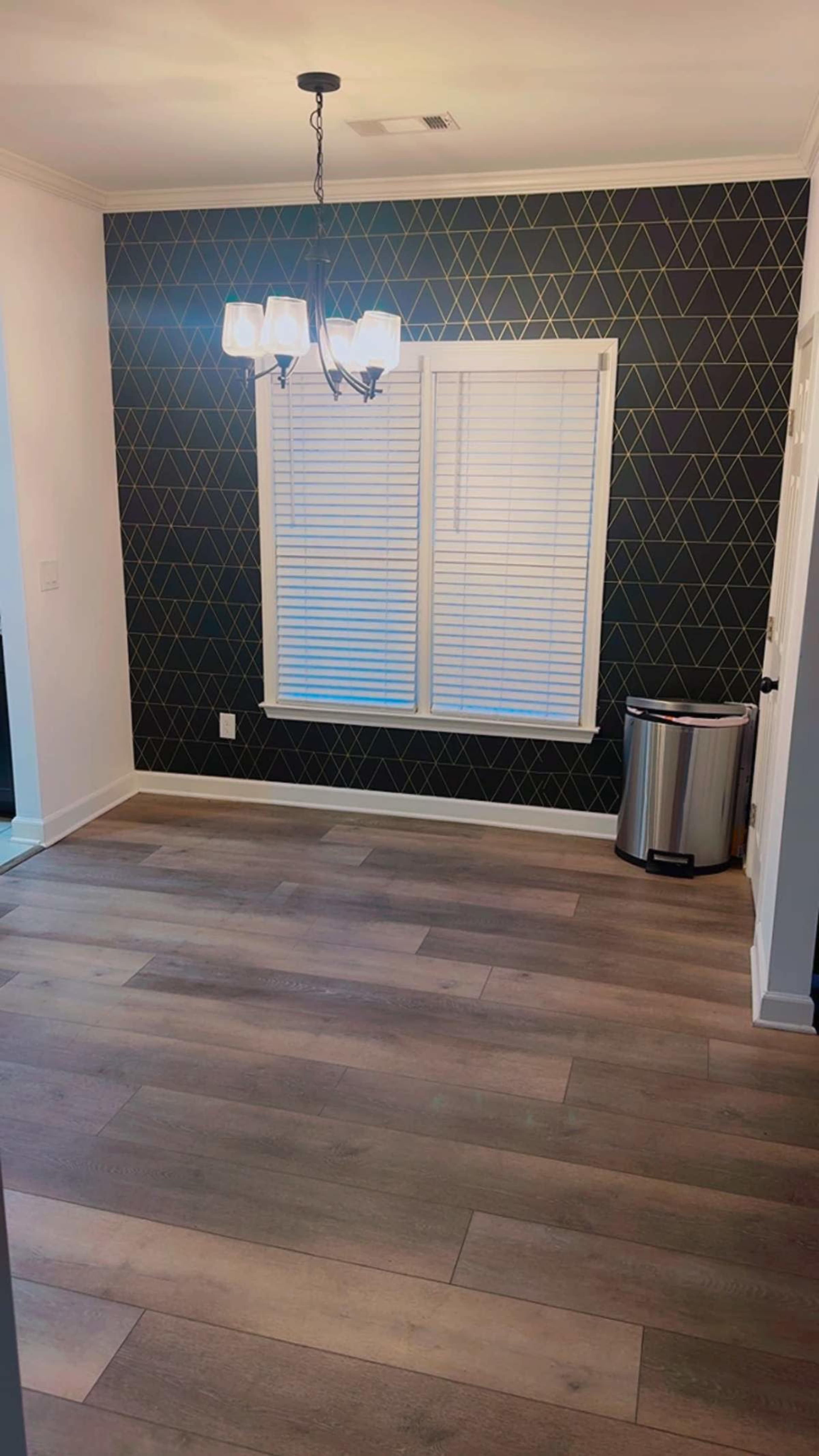 The image shows an empty room with a patterned dark wall, a window covered with white blinds, a chandelier, and a trash can in the corner.