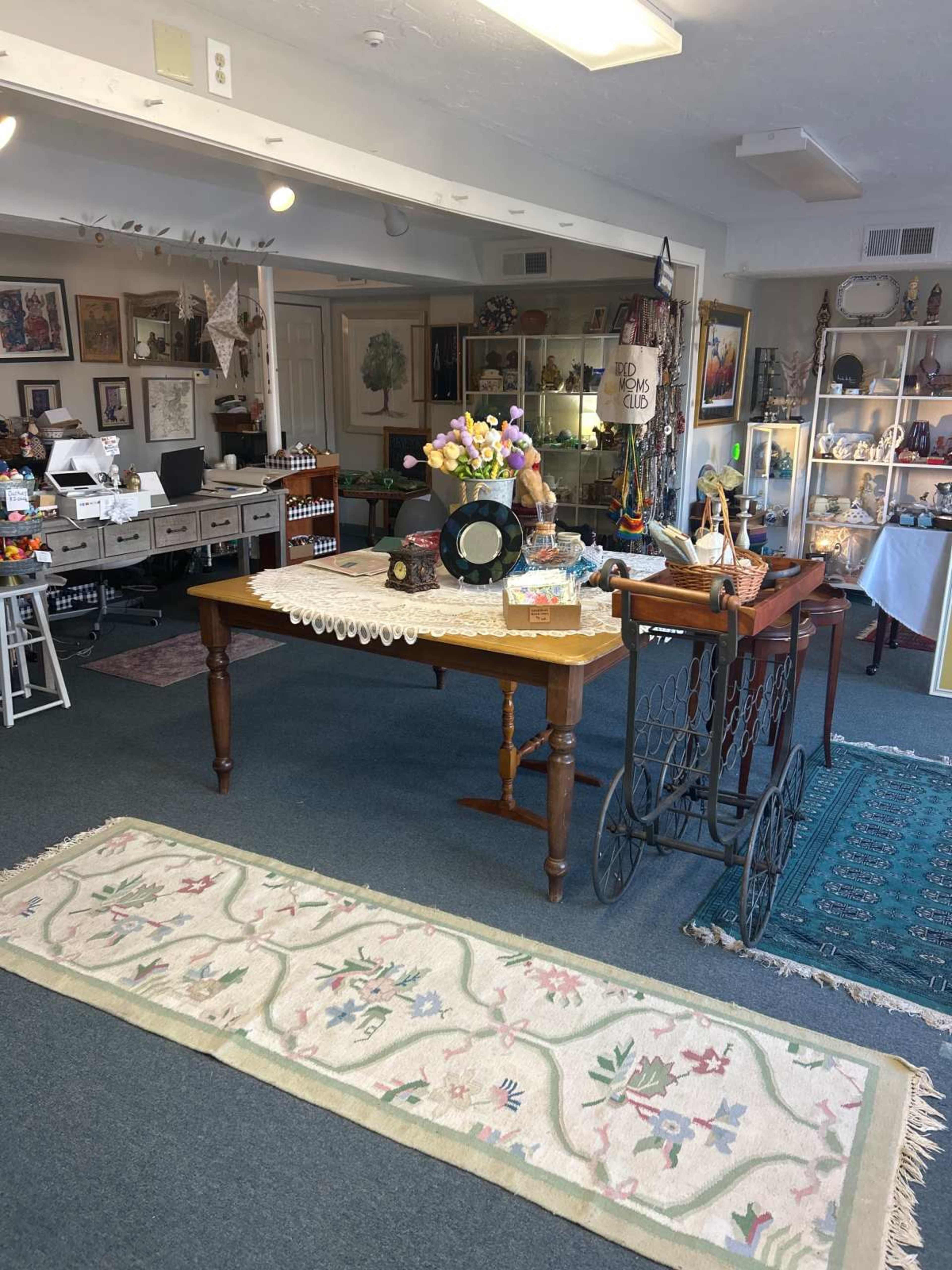 The image shows an interior of a vintage shop featuring two tables, decorative items, and a patterned rug on the floor.