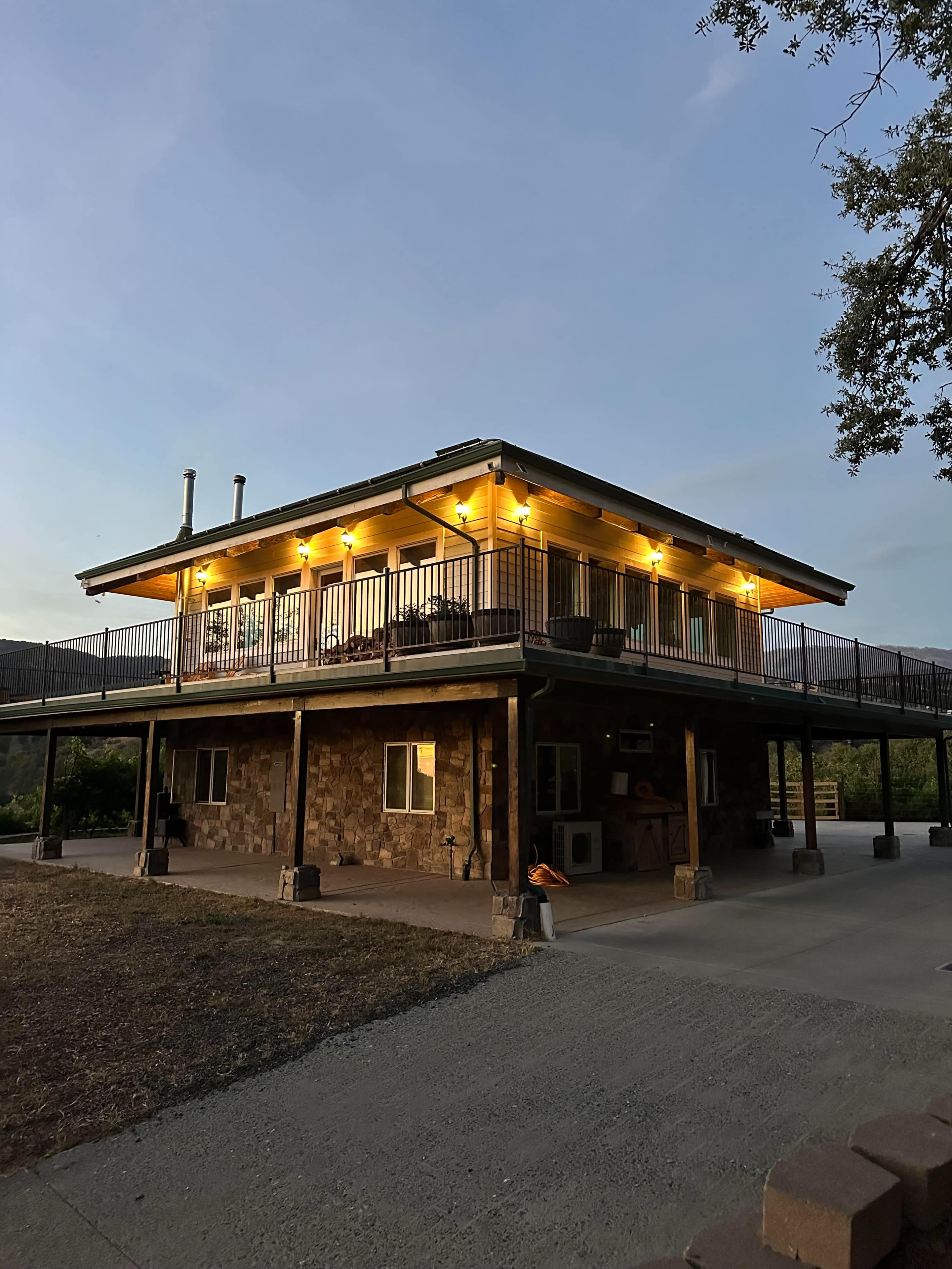 The image shows a two-story house with stone and wooden siding, illuminated by lights, set against a twilight sky.